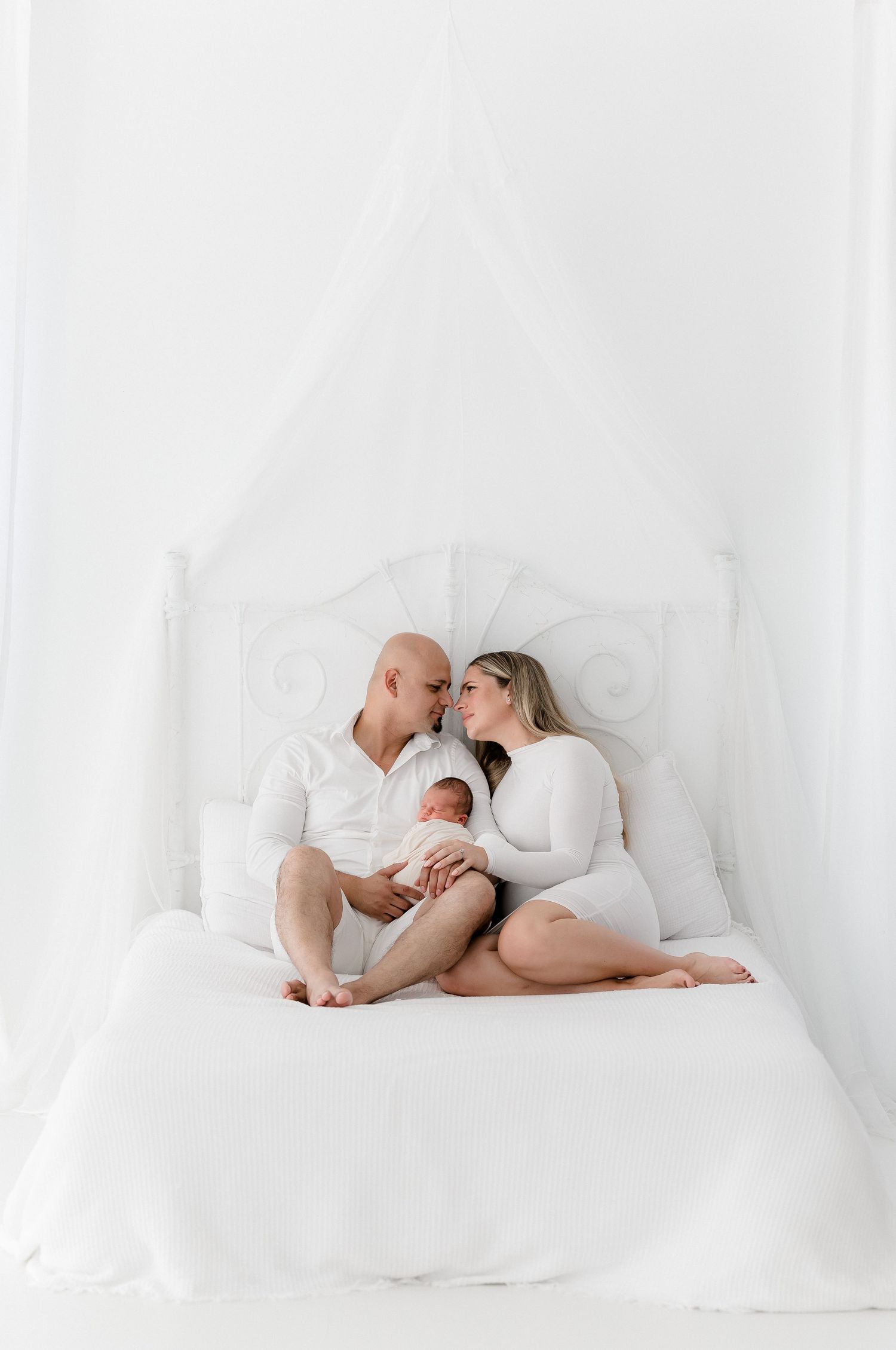 A peaceful family scene with parents and newborn baby resting together on white bedding in soft natural light.