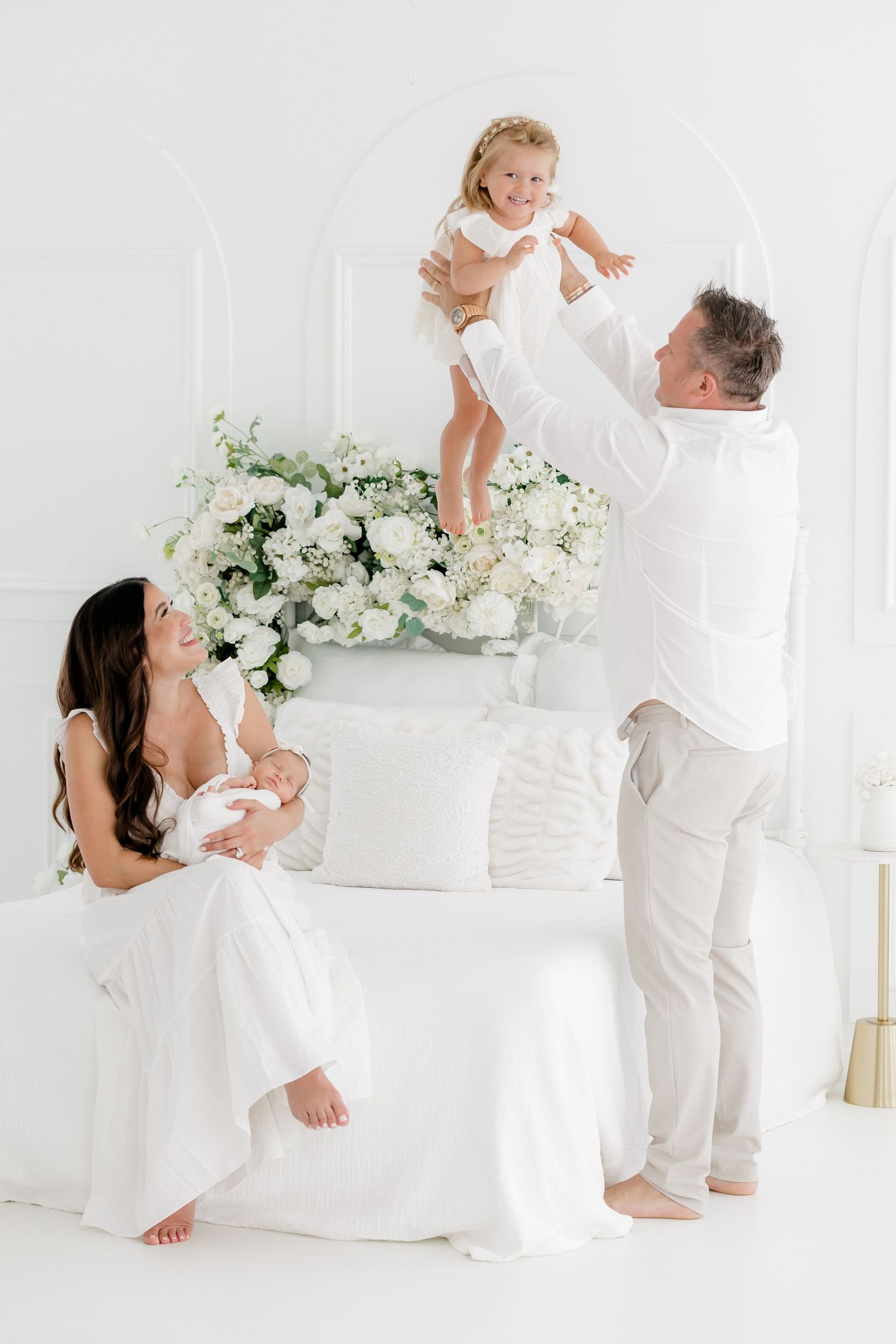 A family wearing white clothes plays together on a bright white couch with floral arrangements in a minimalist setting.