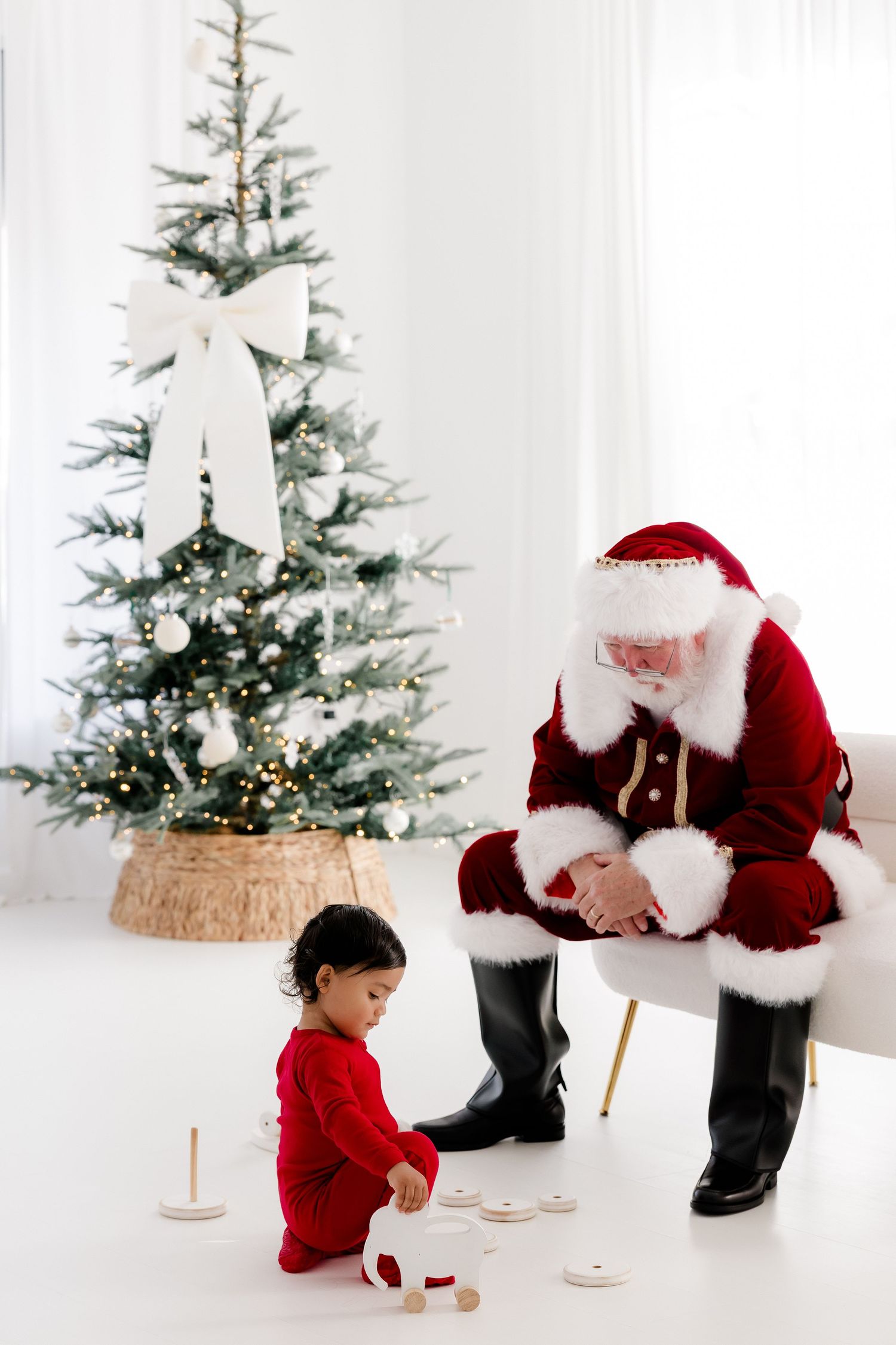 Santa Claus sits beside a toddler in red pajamas playing with toys near a decorated Christmas tree.