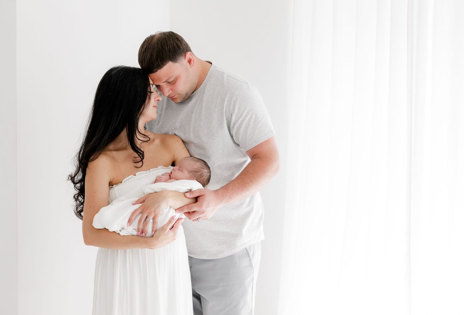 Parents in white clothing hold and gaze lovingly at their newborn baby against a bright white backdrop.