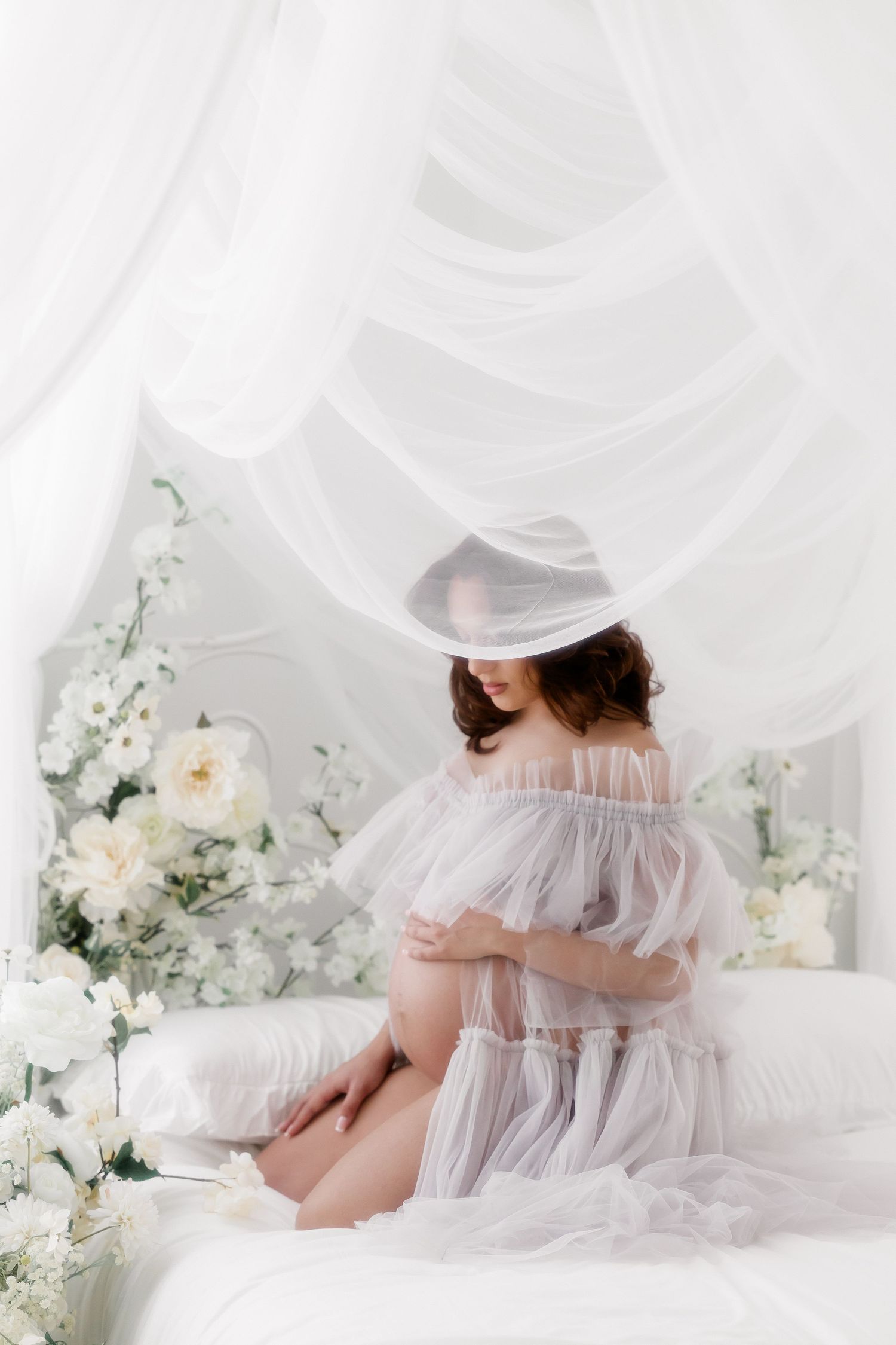 Ethereal maternity photo with flowing sheer white fabric, white roses and delicate floral arrangements in dreamy setting.