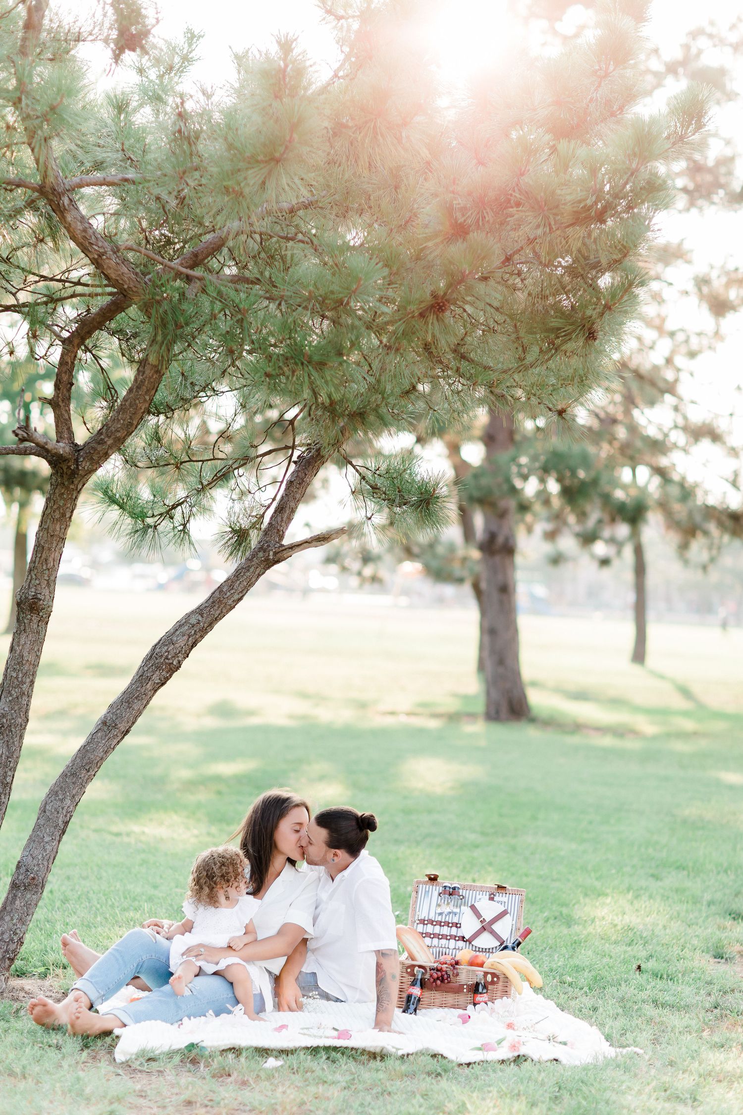 Family lounging together on white blanket for outdoor photoshoot in dreamy sunlit park setting with pine trees.