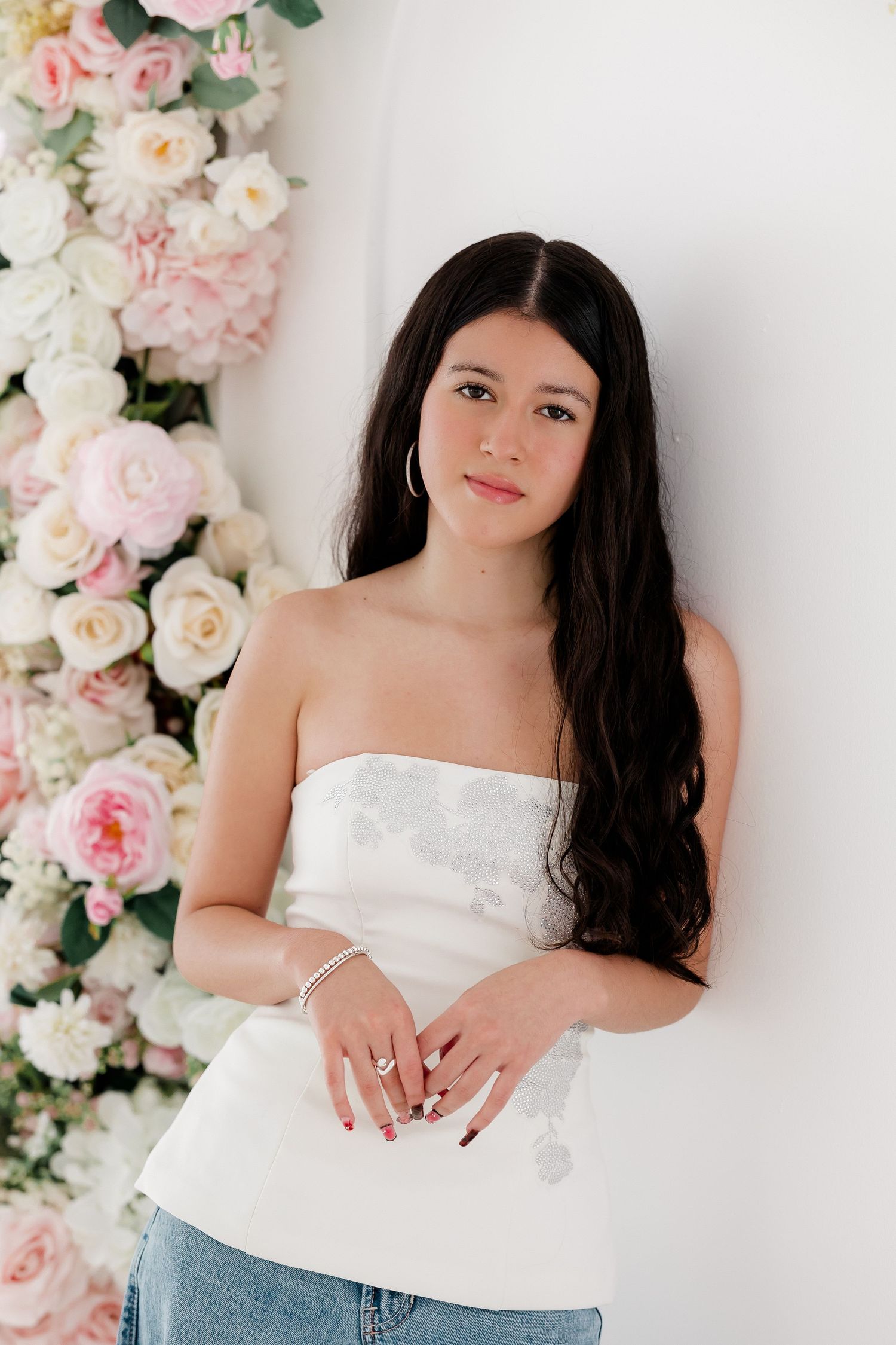 Portrait against white wall with pink and white rose floral arrangement, wearing strapless white top and denim.