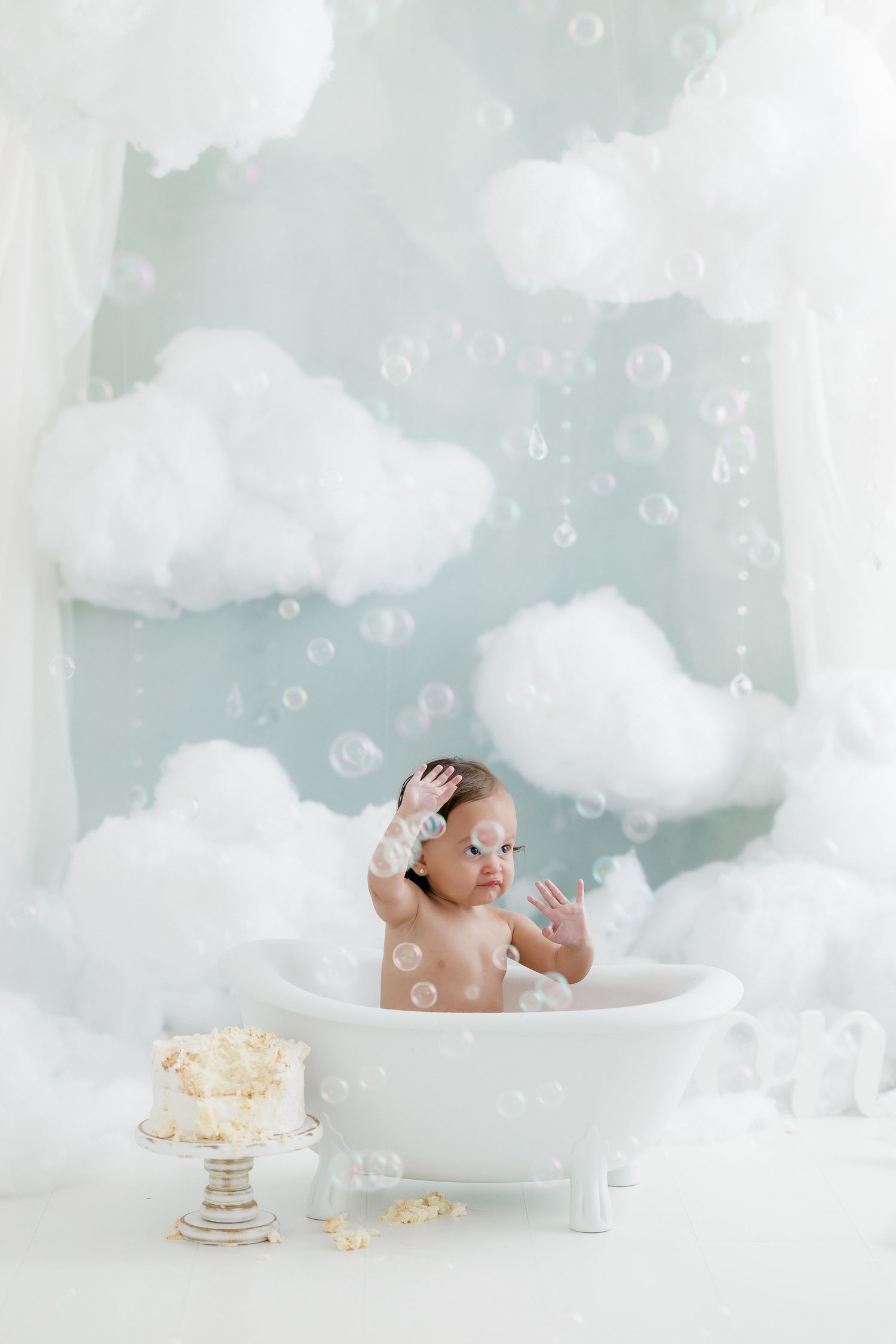 Baby in white bathtub with soap bubbles and cloud decorations during a first birthday cake smash photo session.