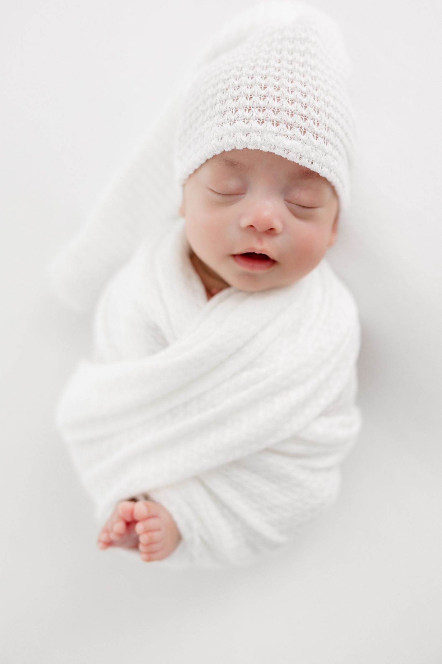Newborn baby swaddled in white blanket wearing knit hat against white background.