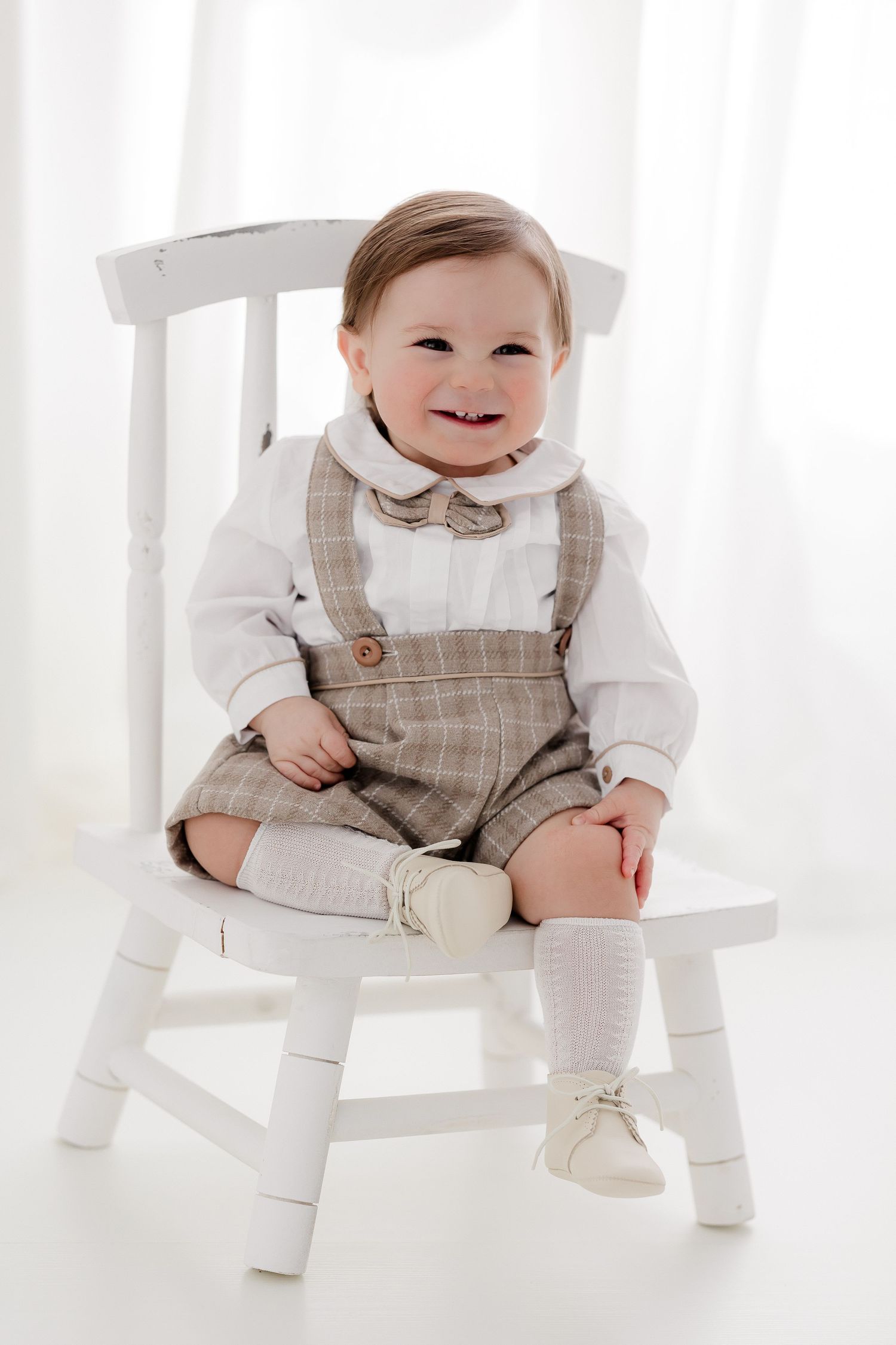 Baby in vintage-style tan overalls and bow tie sits smiling on a white wooden chair.