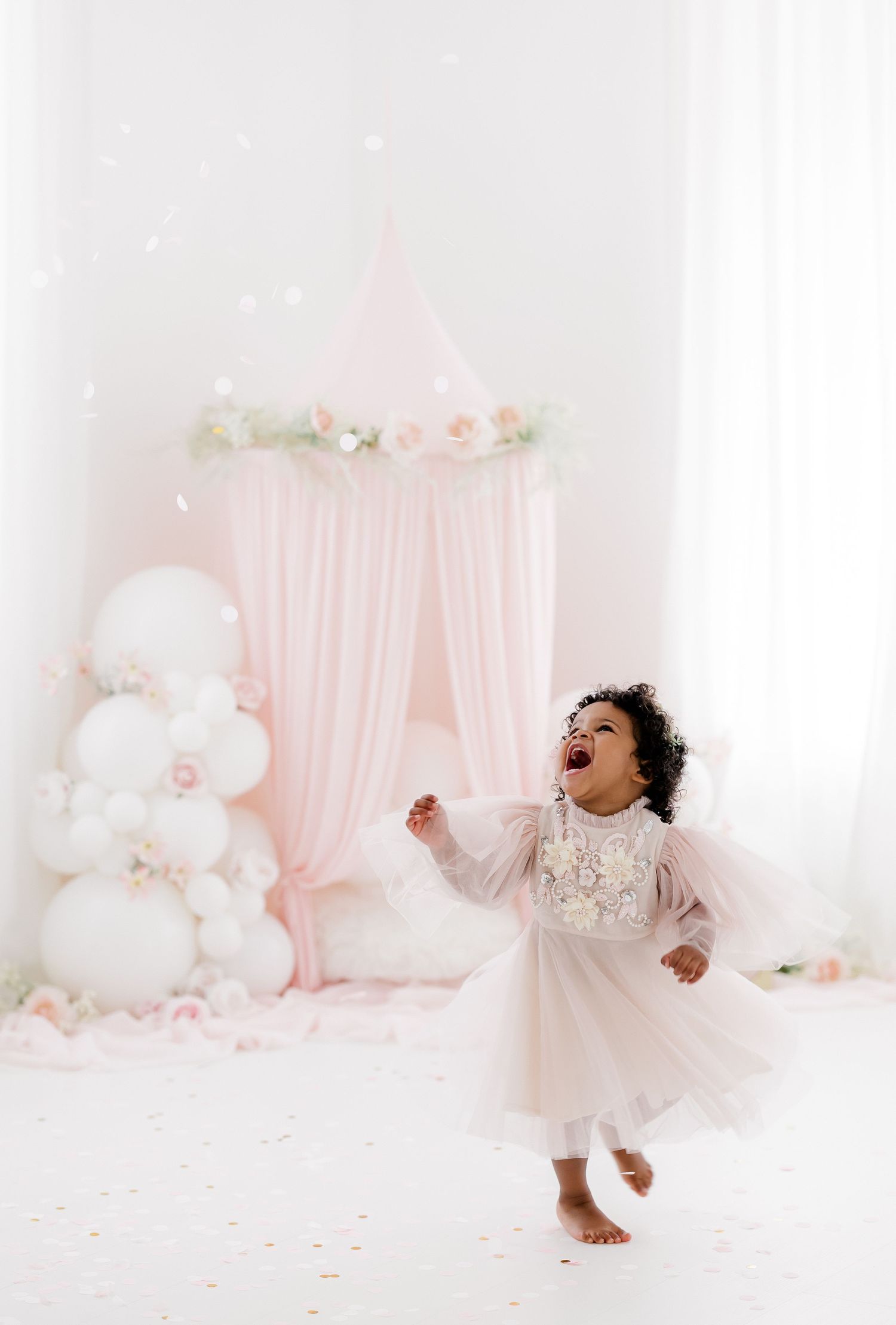 Joyful toddler in white dress dancing against pink and white balloon backdrop.