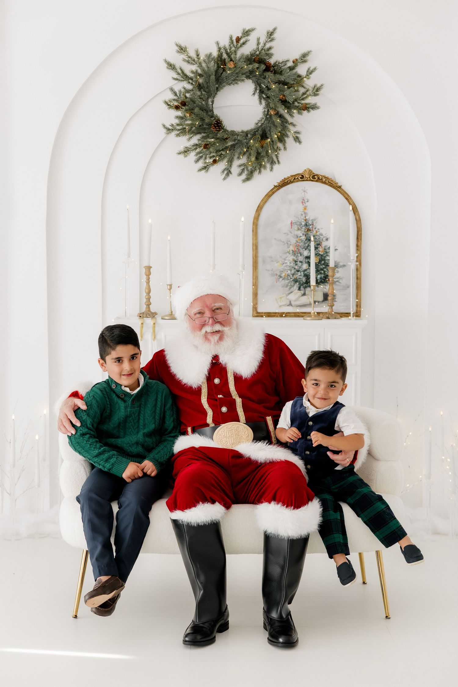 Two boys sit with Santa Claus on a white chair in a festive holiday studio setting with wreath and fireplace.