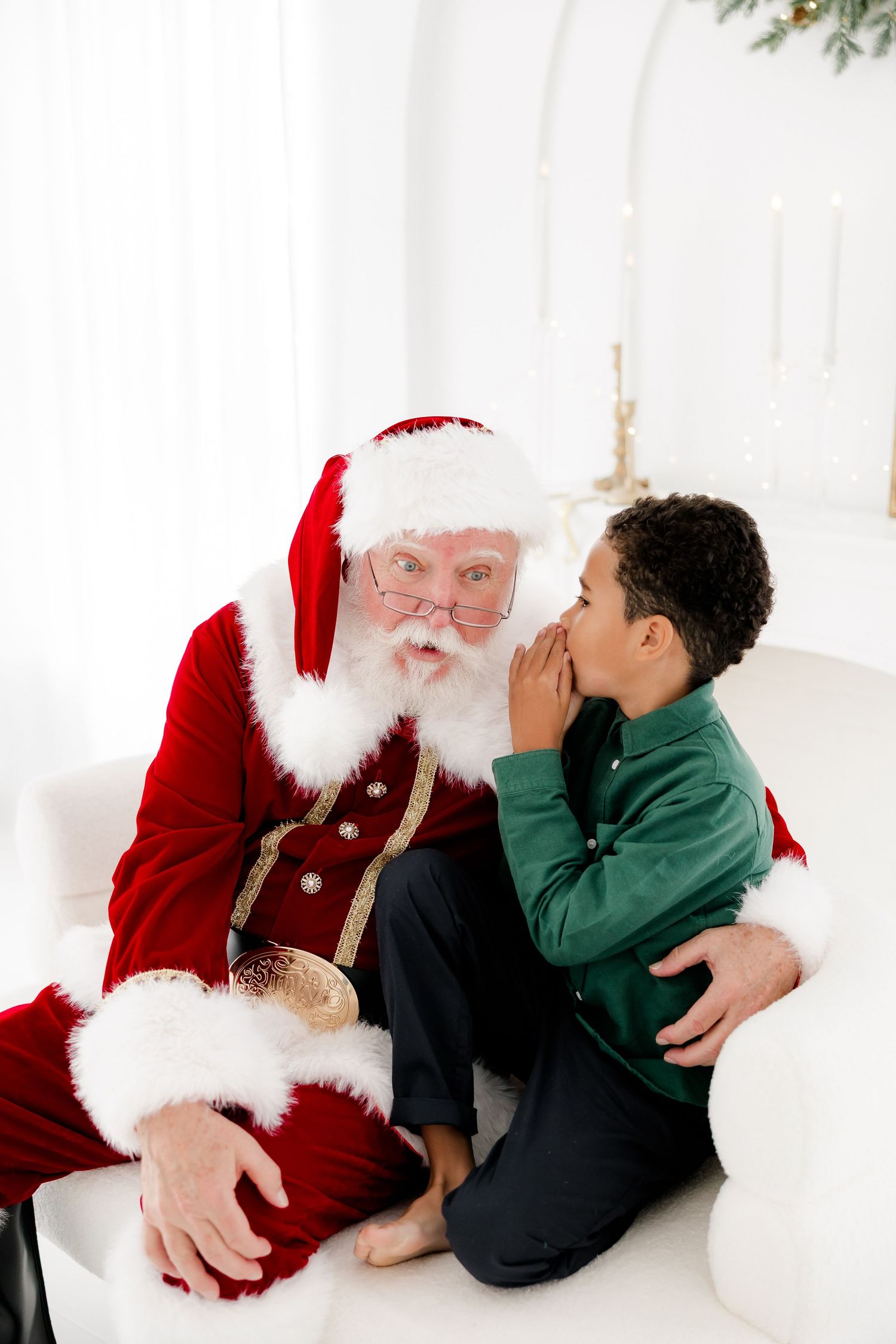 A young boy whispers his Christmas wishes to Santa Claus while sitting together on a white sofa.