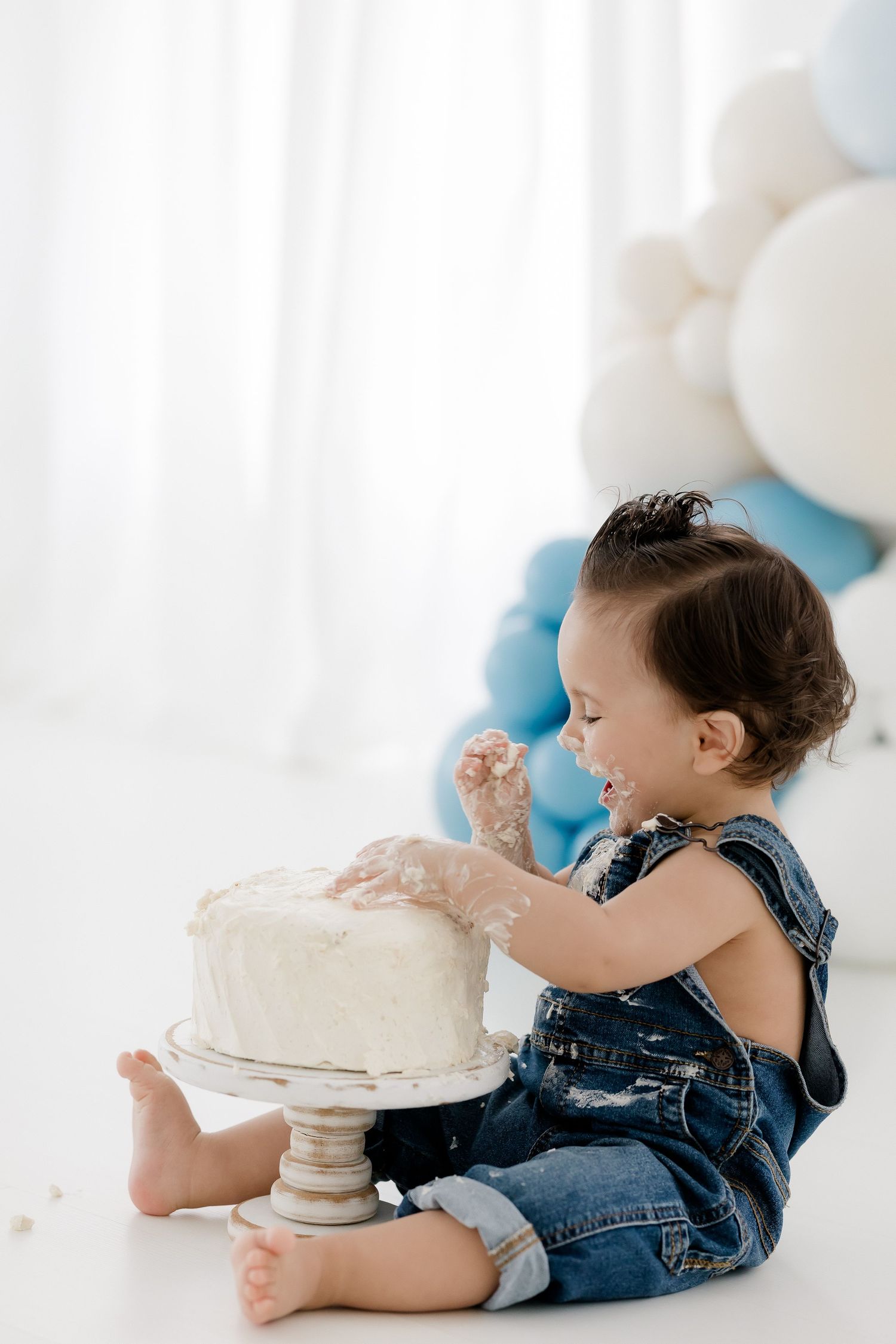 A baby in denim overalls lying on the floor reaching for a white frosted cake on a pedestal.