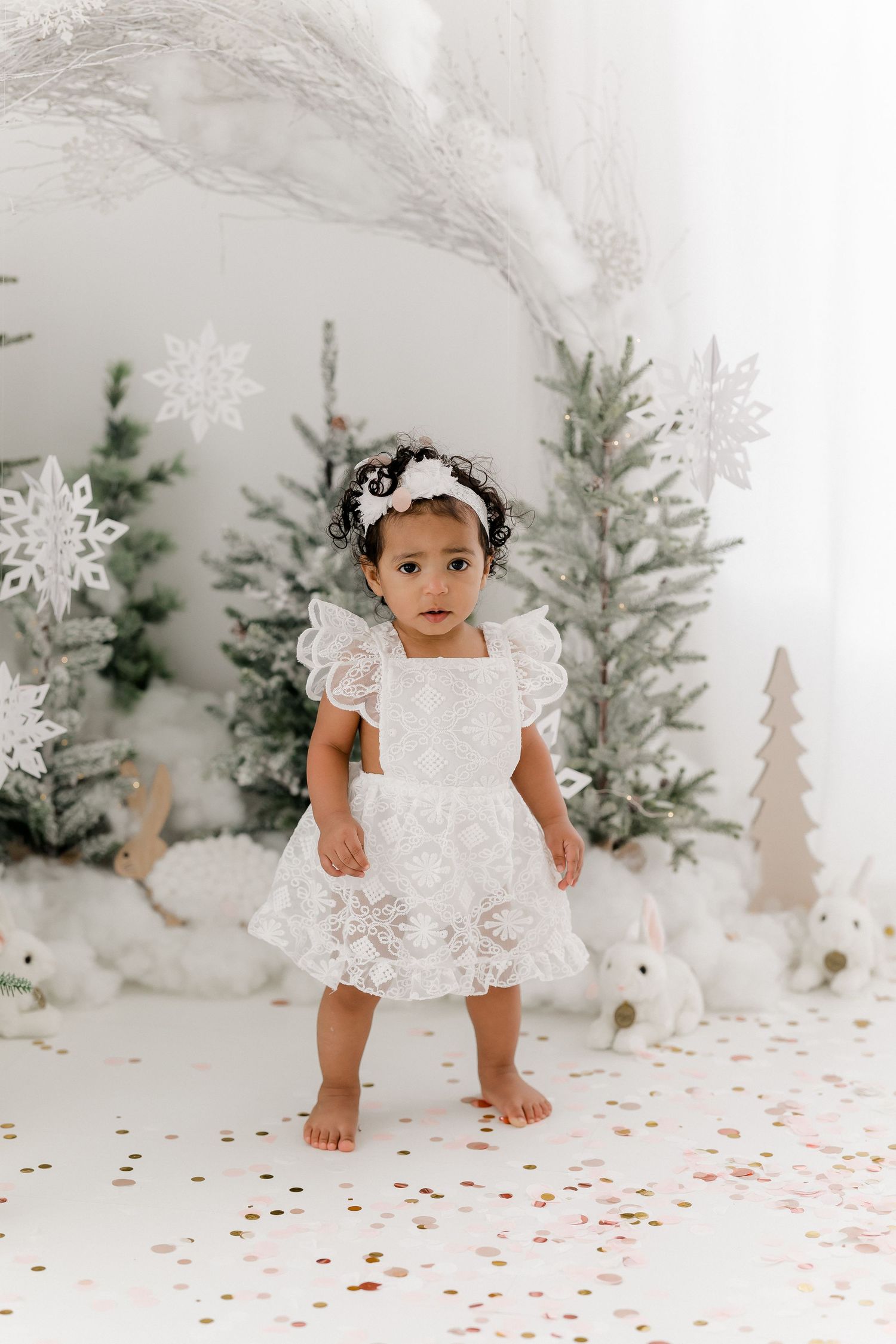 Whimsical winter photo shoot featuring a white lace dress against snowy evergreen trees and scattered confetti.
