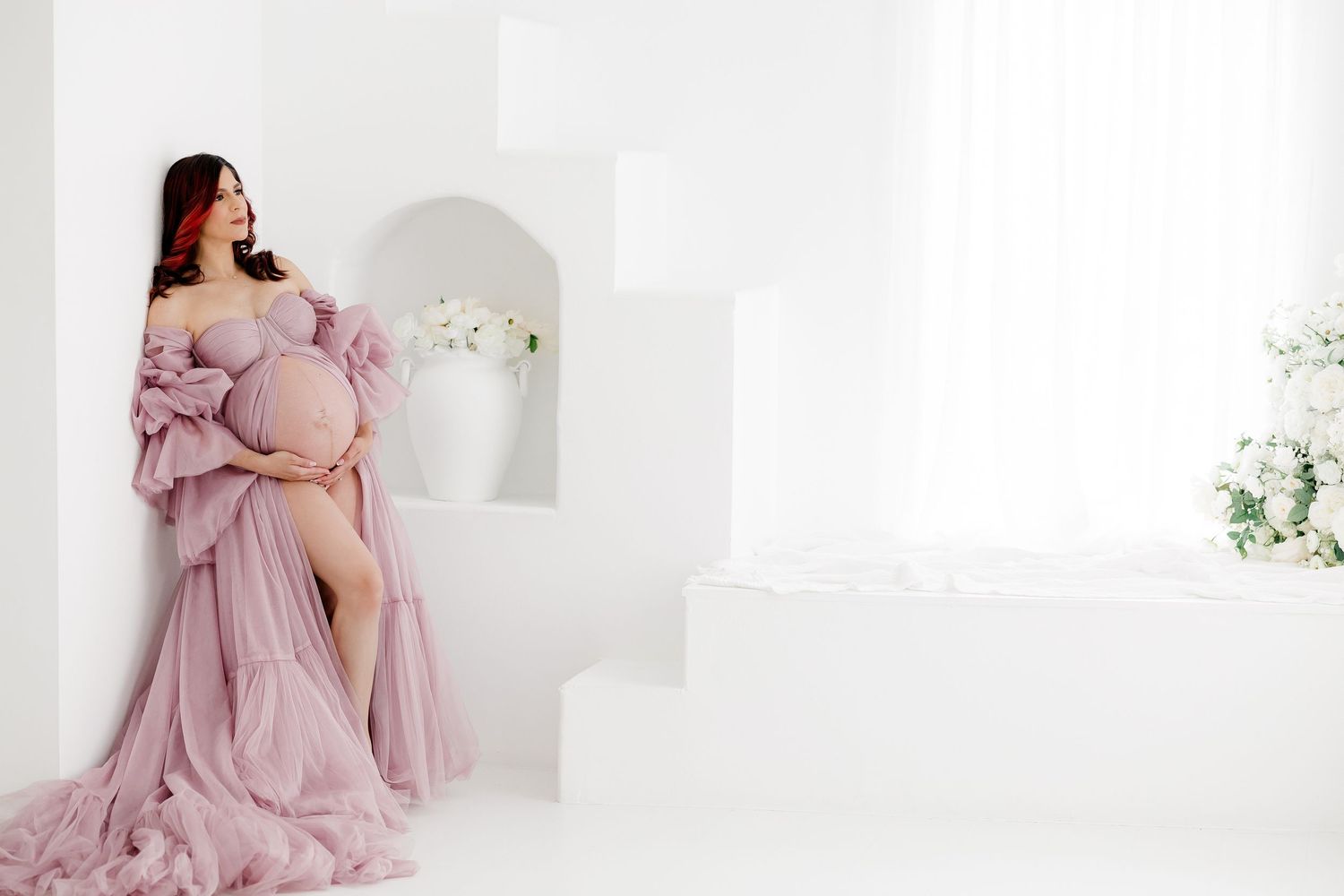 Soft pink maternity portraits with woman in flowing dress against white background.