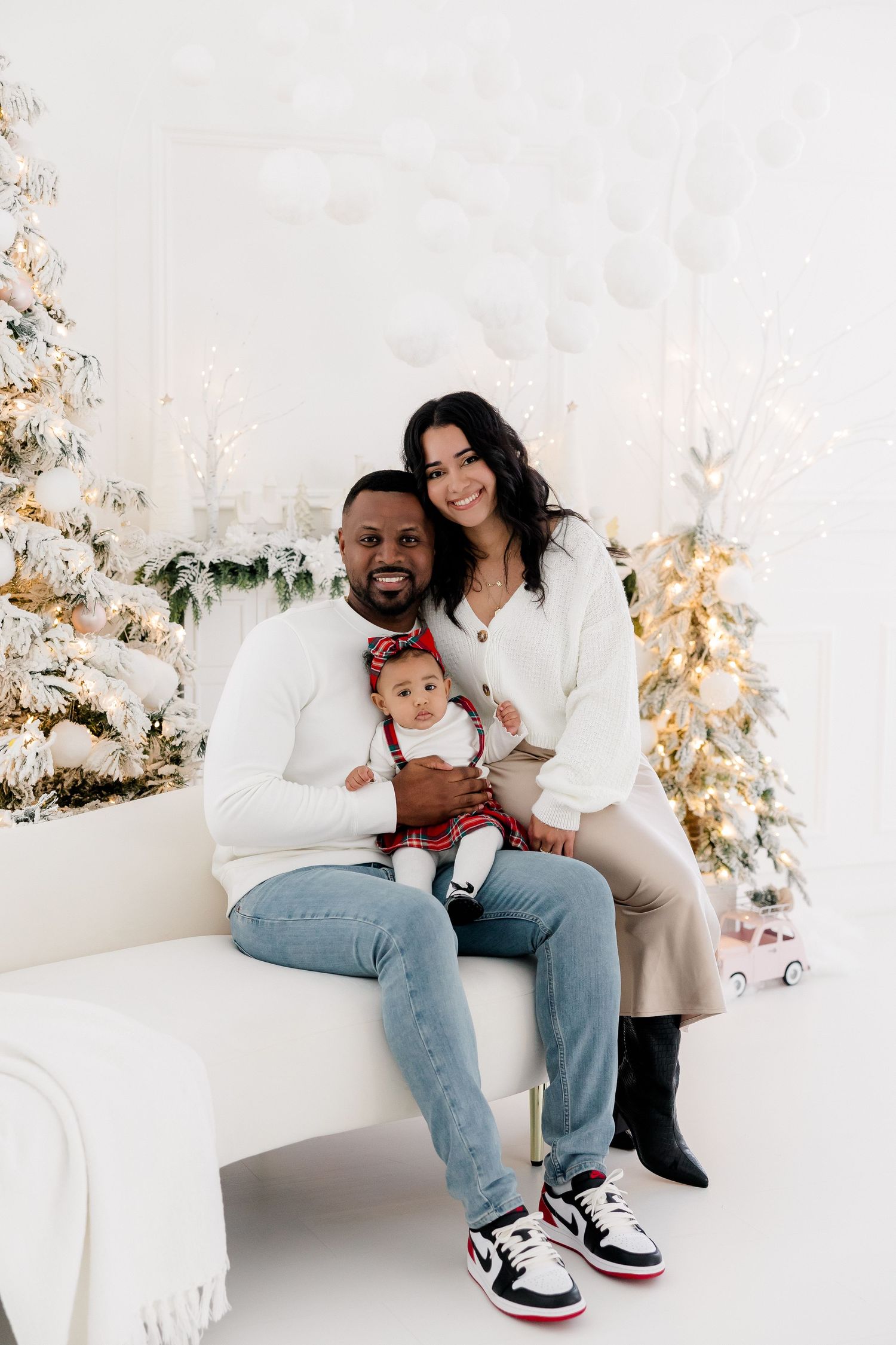 Family relaxes on white couch surrounded by decorated Christmas trees in modern studio setting.