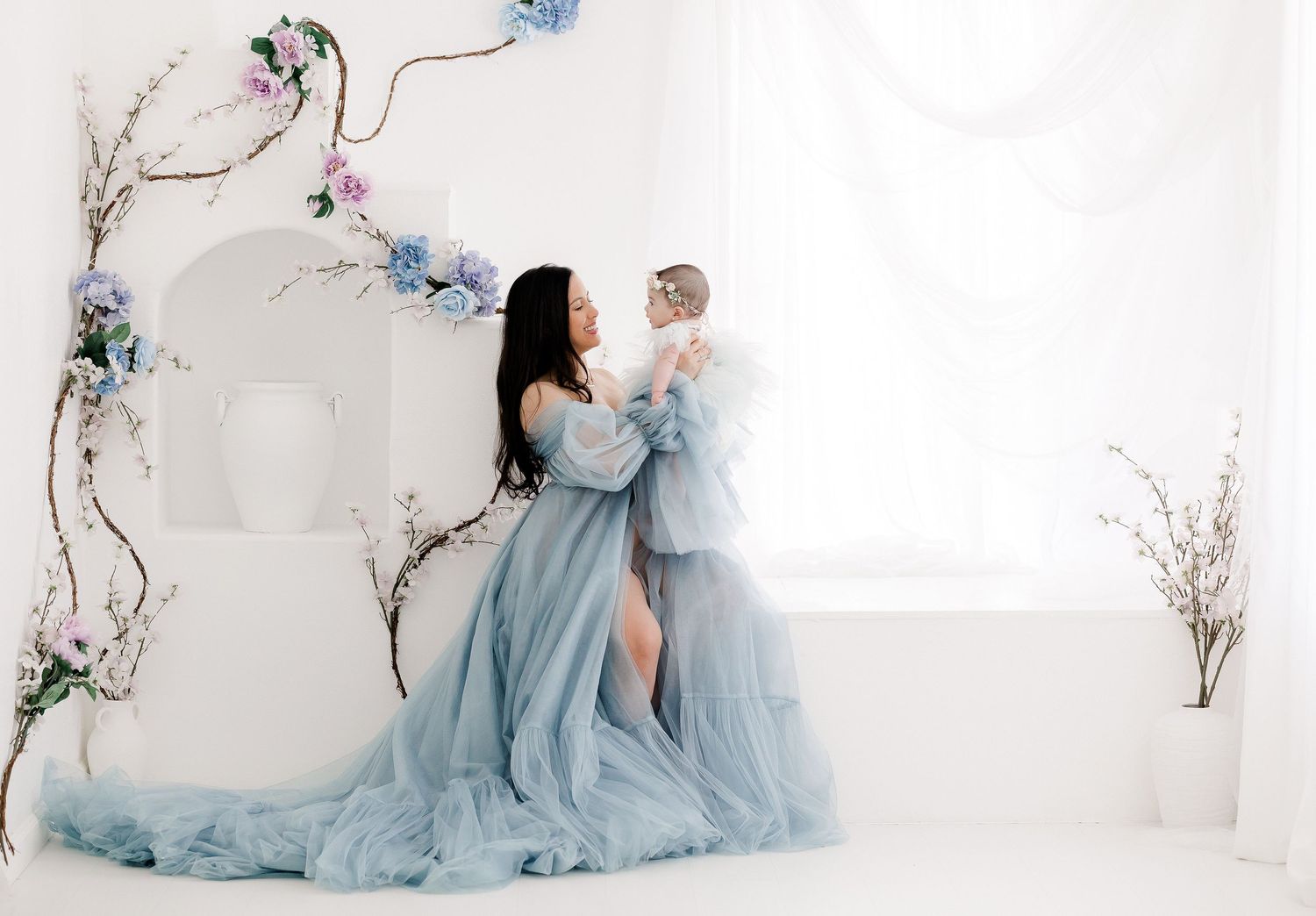 A dreamy portrait in a white room with floral decor features flowing powder blue gowns and delicate spring branches.