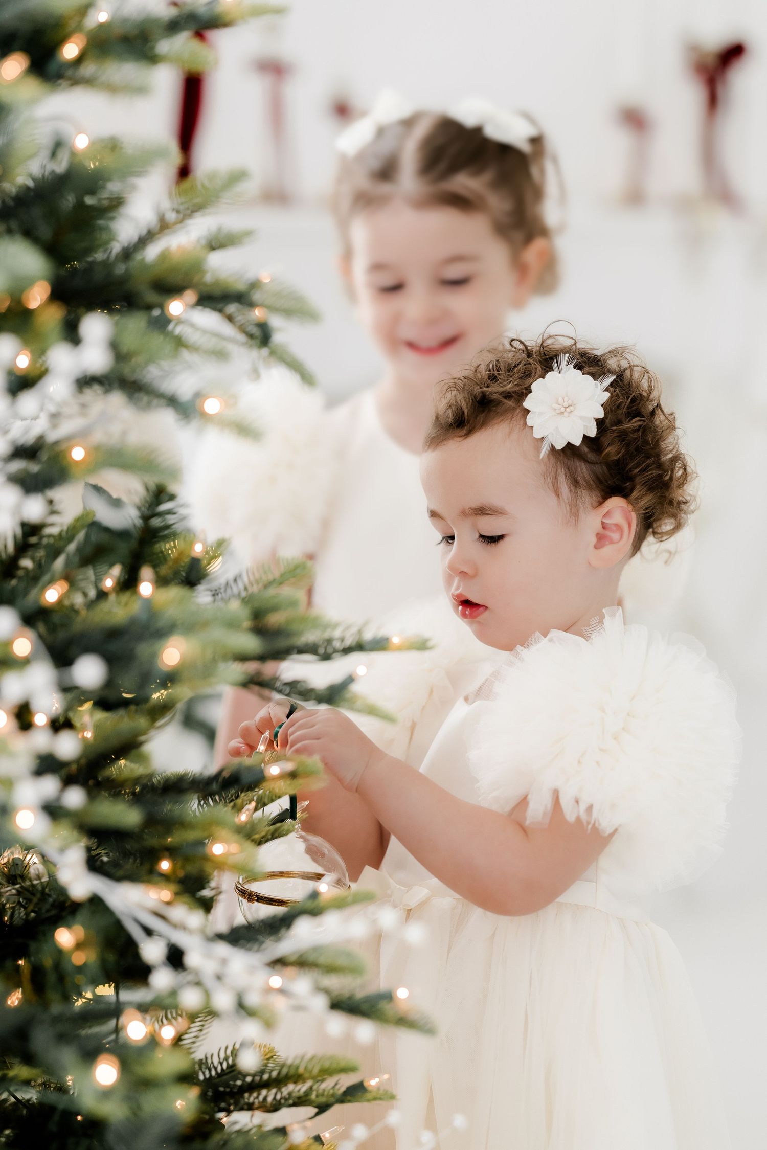 Two young girls in white dresses decorate a Christmas tree with lights and ornaments in a bright holiday setting.