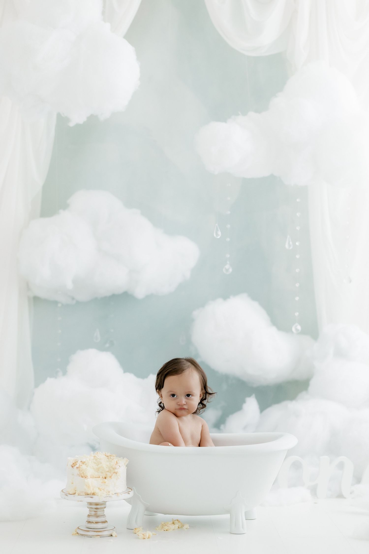 Adorable baby sits in white clawfoot bathtub surrounded by fluffy clouds in soft blue studio.