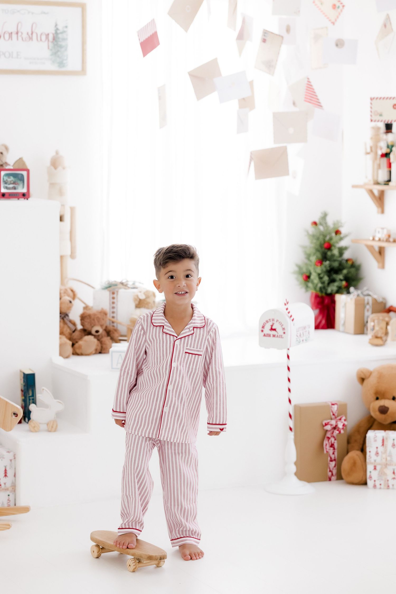 Boy in red striped Christmas pajamas standing on a wooden skateboard in a festive white holiday room with gifts.