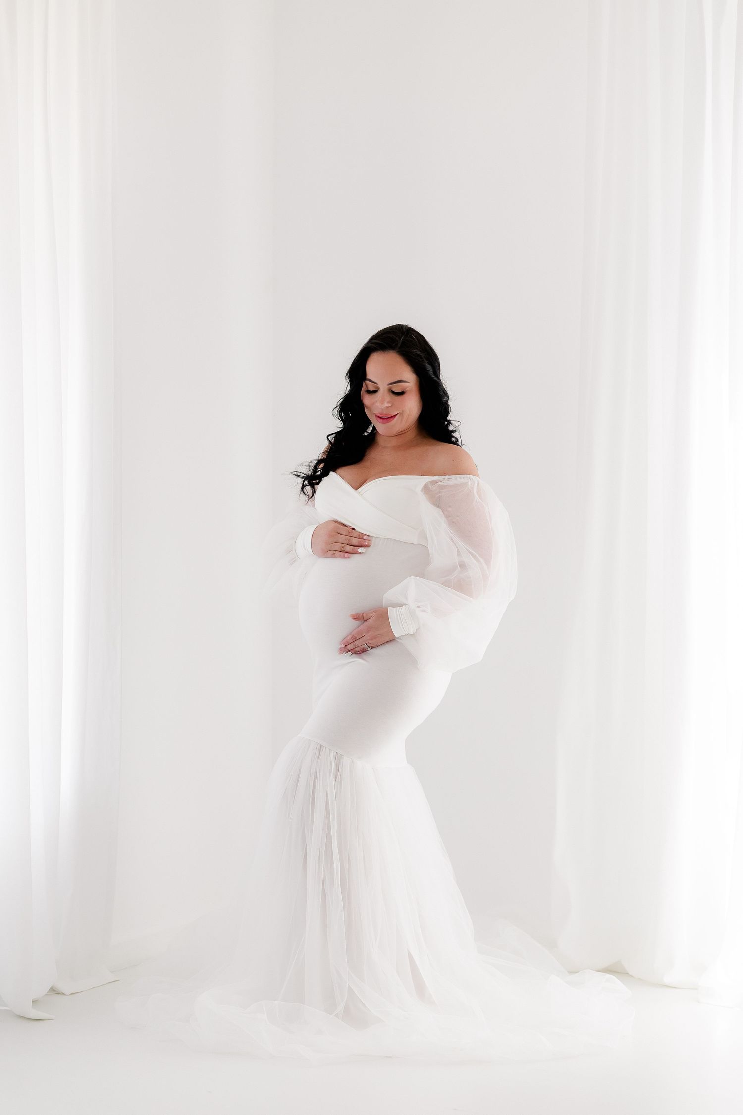 Classic maternity photography in white flowing dress against bright studio backdrop.
