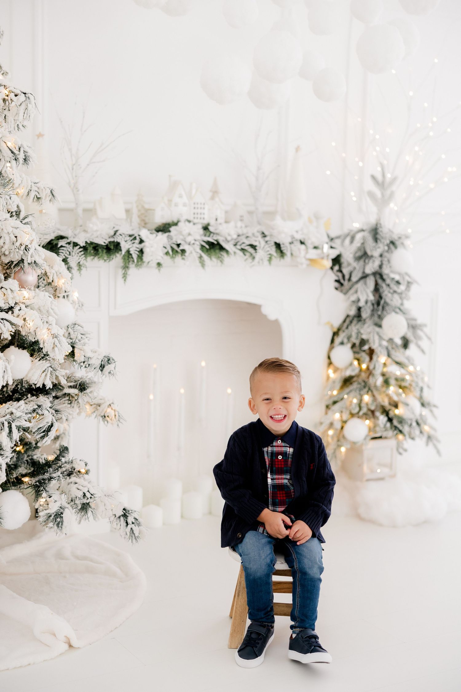 Child poses in winter wonderland setting with snow-covered trees and holiday decorations in bright white studio.