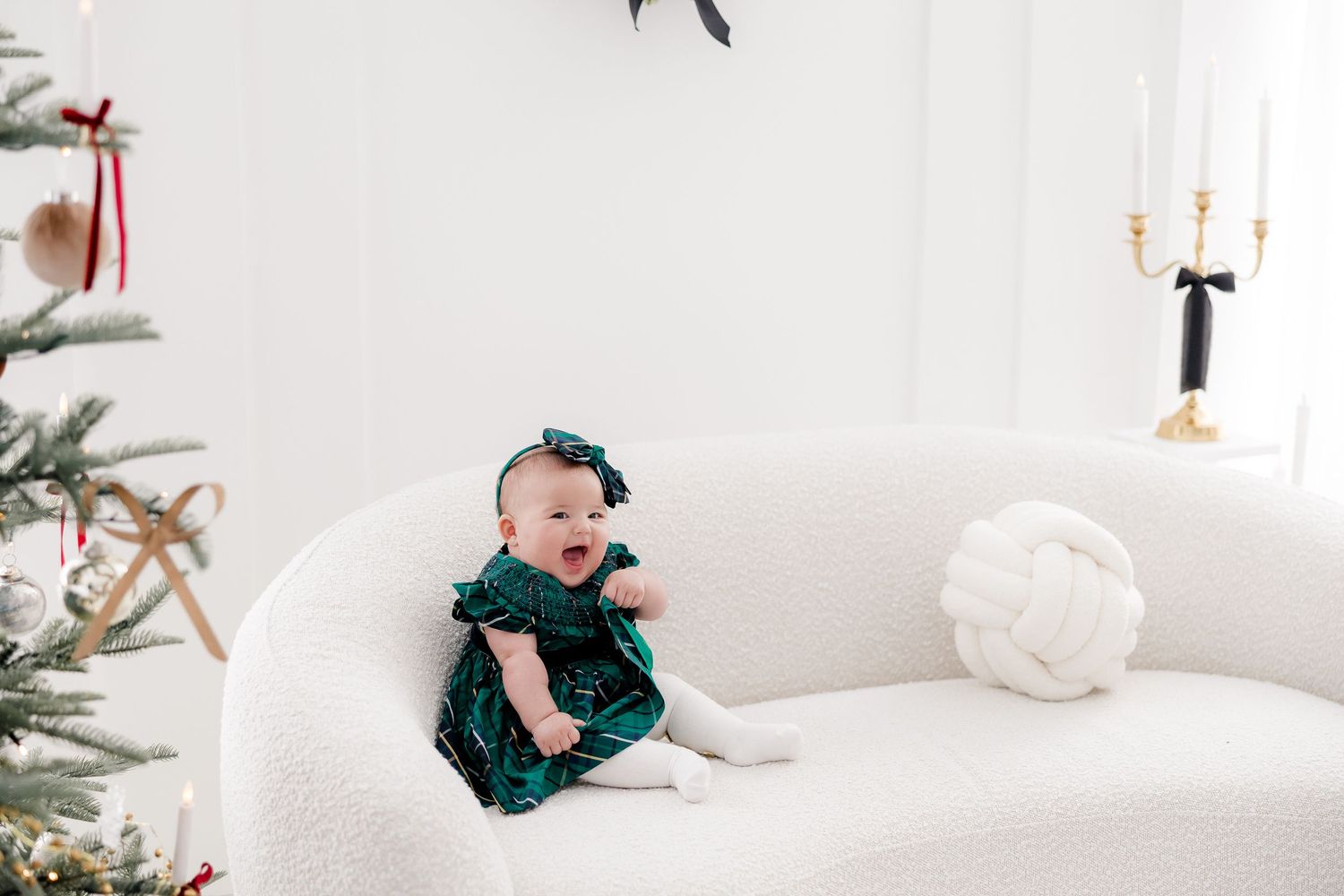 Baby wearing green velvet dress sits on white couch near decorated Christmas tree.