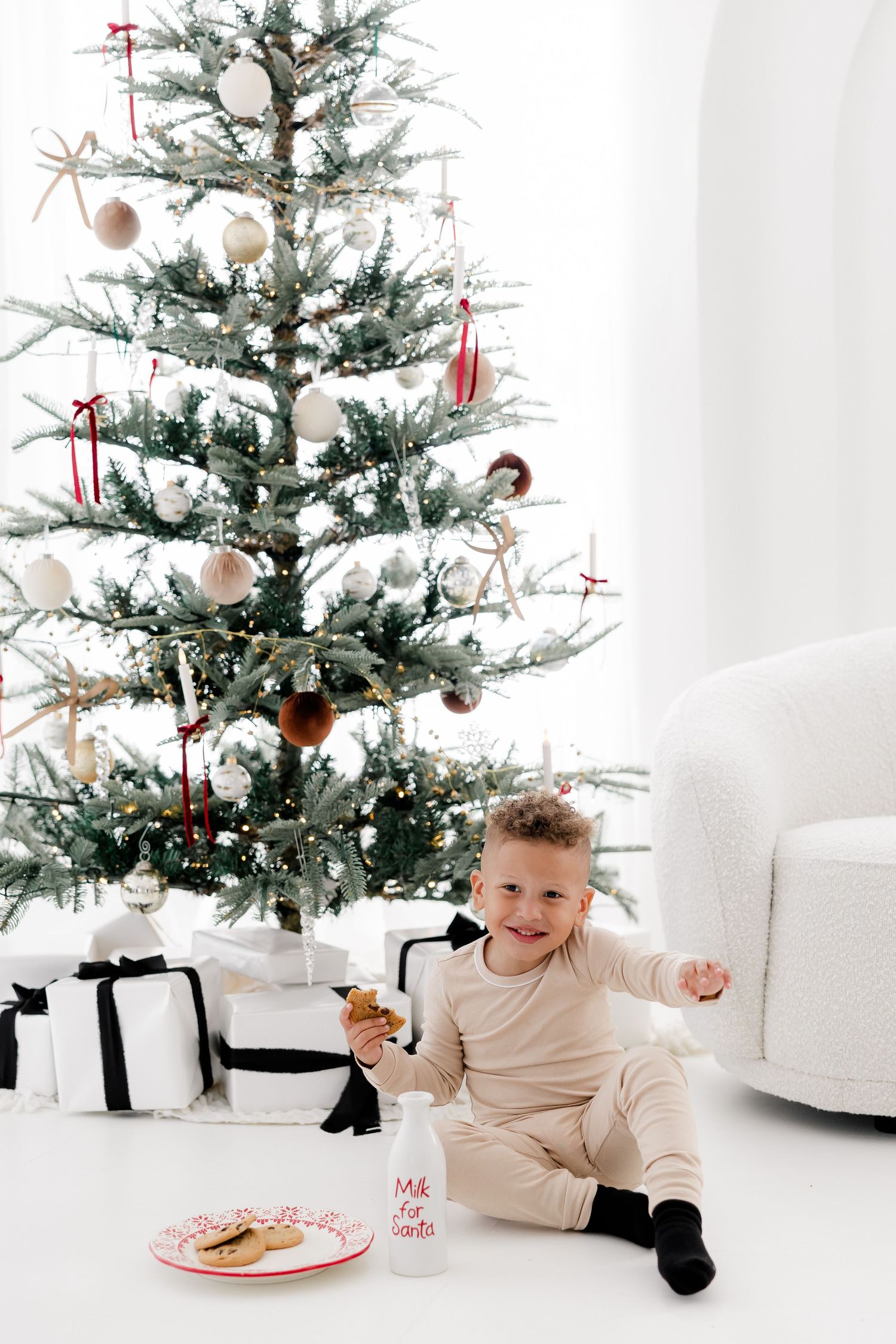 Child enjoying Christmas morning treats next to decorated tree in white studio setting.