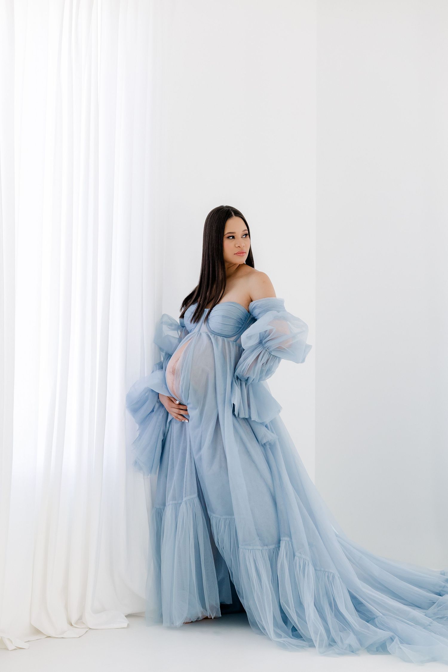 Ethereal maternity photo shoot featuring a flowing powder blue tulle gown against a white backdrop.