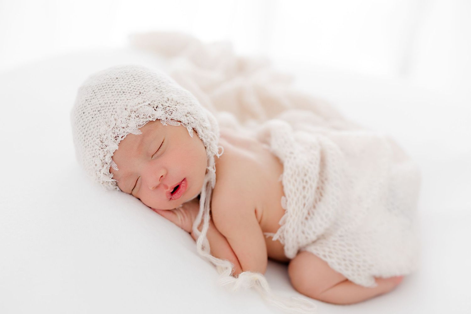 Peaceful newborn sleeping in white lace wrap and bonnet on light background.