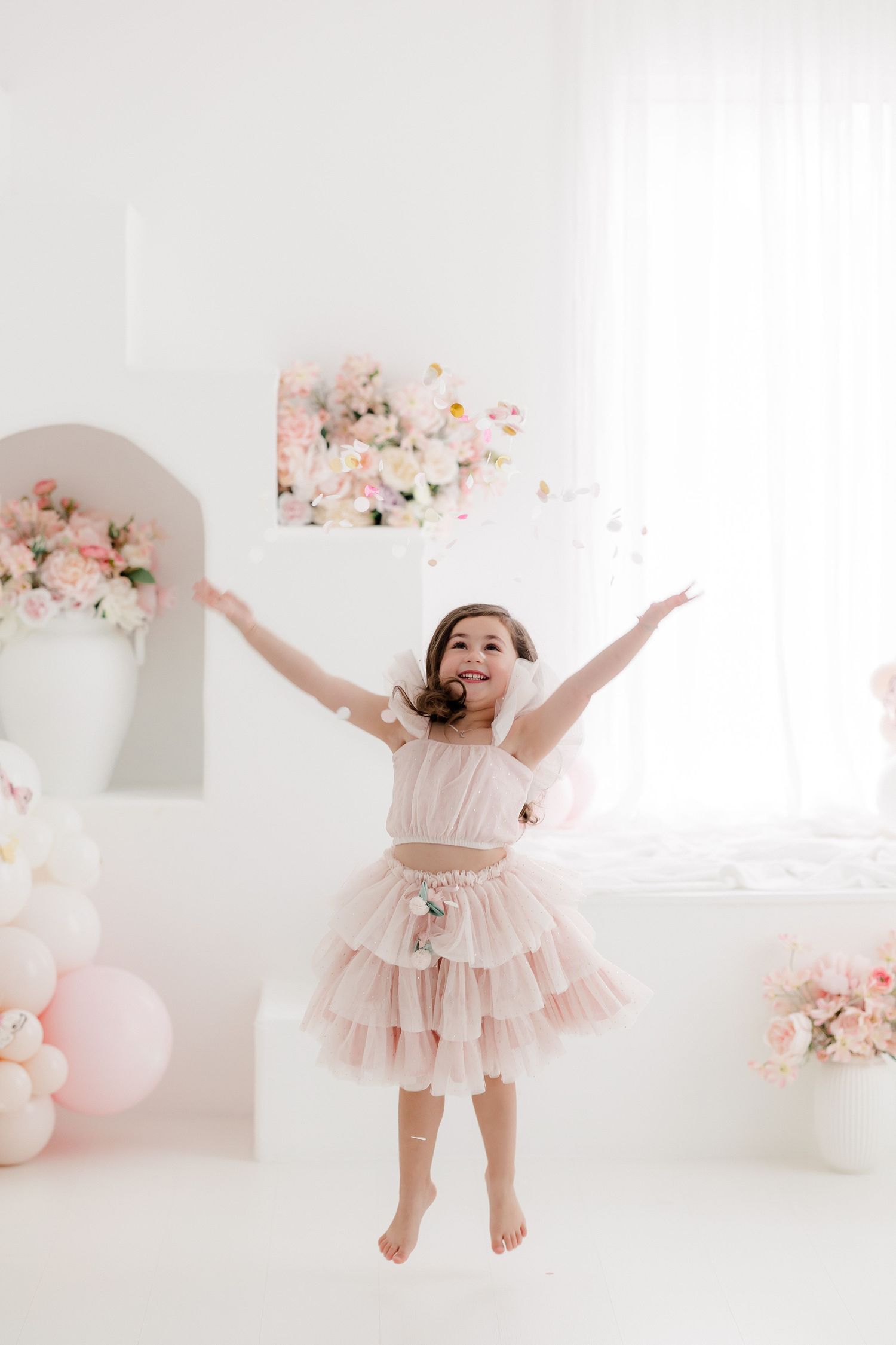 A child in a pink tulle dress twirls and dances in a bright white studio with floral decorations and balloons.