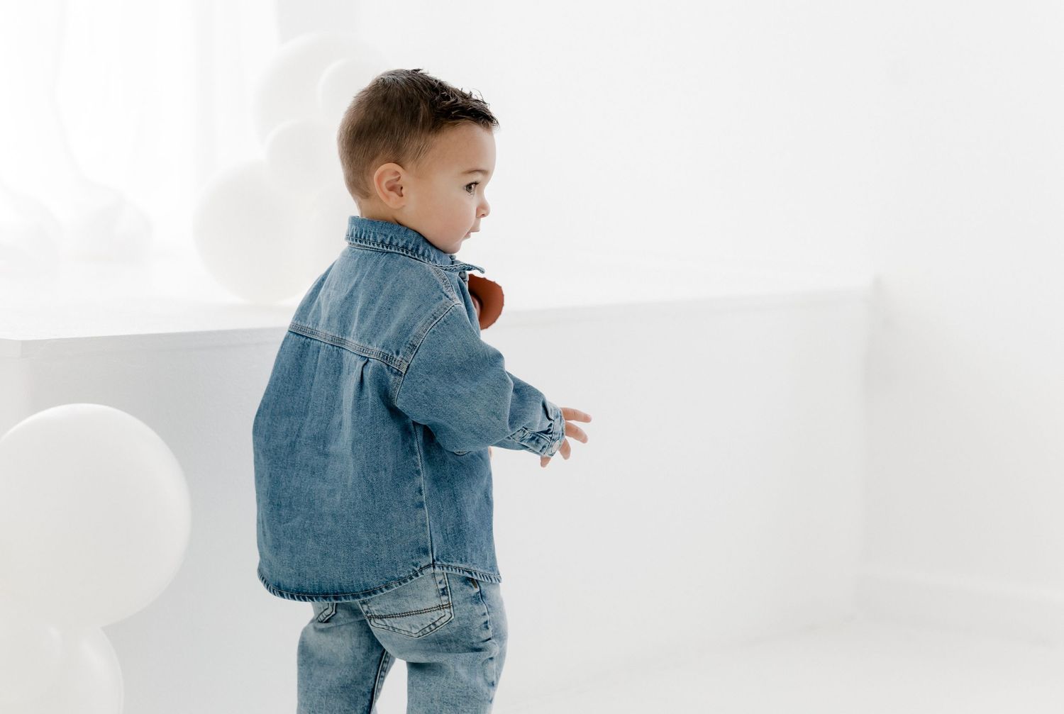 A toddler in a denim jacket and pants plays while walking across a white background.