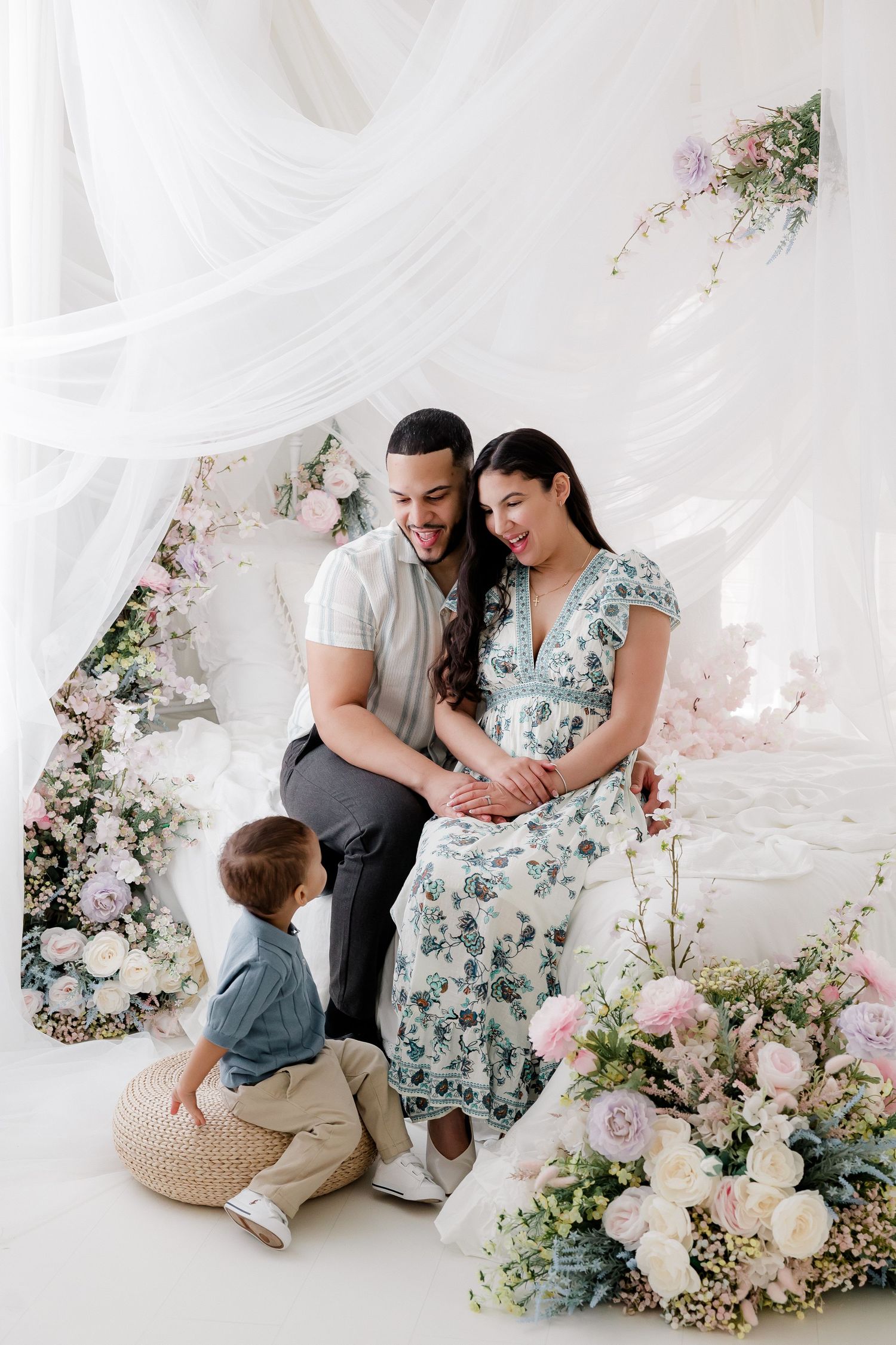 Family maternity photo with floral arch backdrop featuring white draping and pink and white roses in a dreamy studio setting.