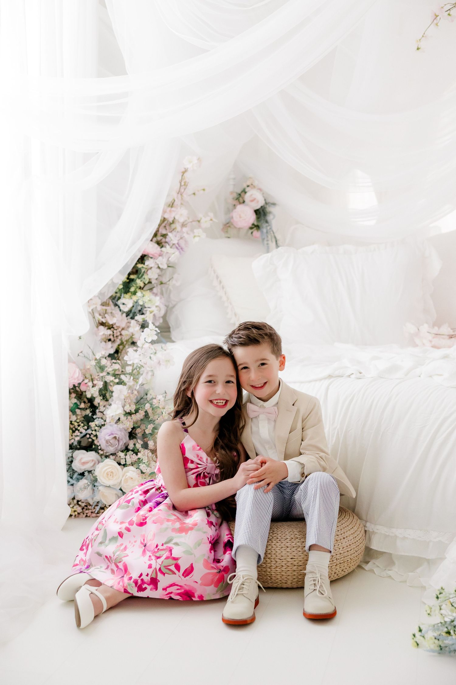 A couple in pink and beige outfits pose together on white fabric with floral decorations in a romantic photoshoot setting.