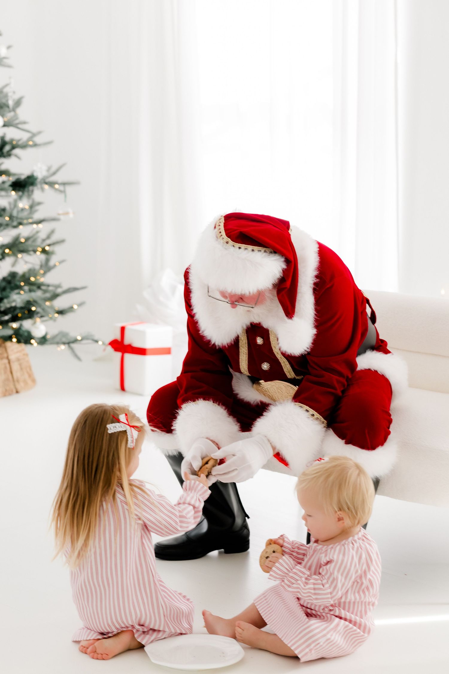 Two children in pink pajamas sit with Santa Claus near a decorated Christmas tree sharing holiday cookies.