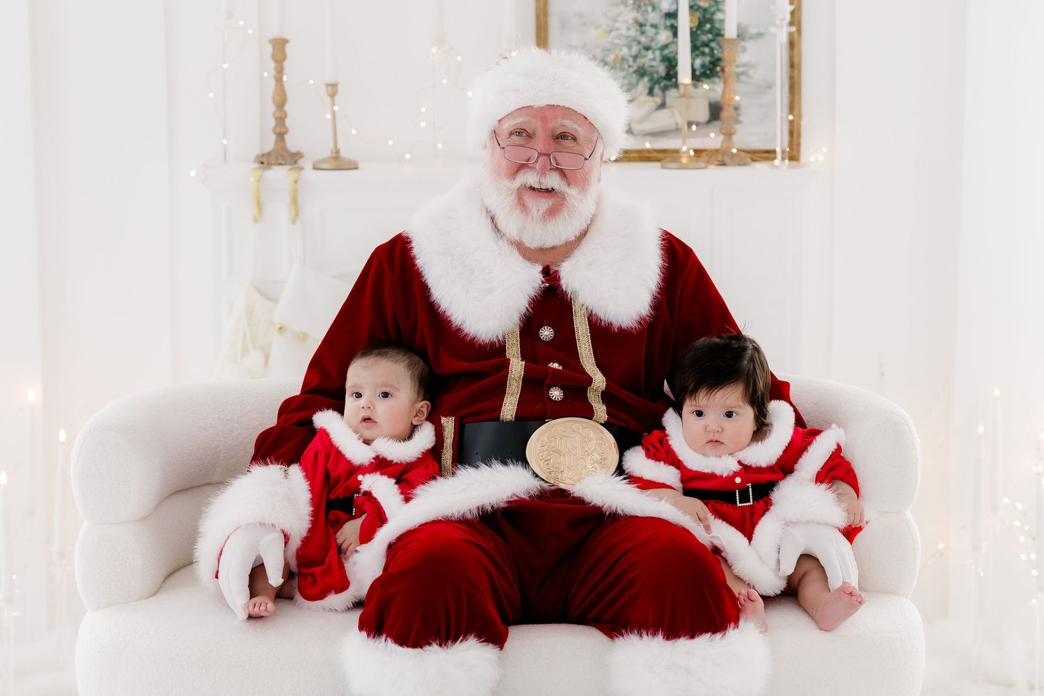 Two babies in Santa outfits sit with a jolly Santa Claus on a white couch in a festive holiday photo setting.