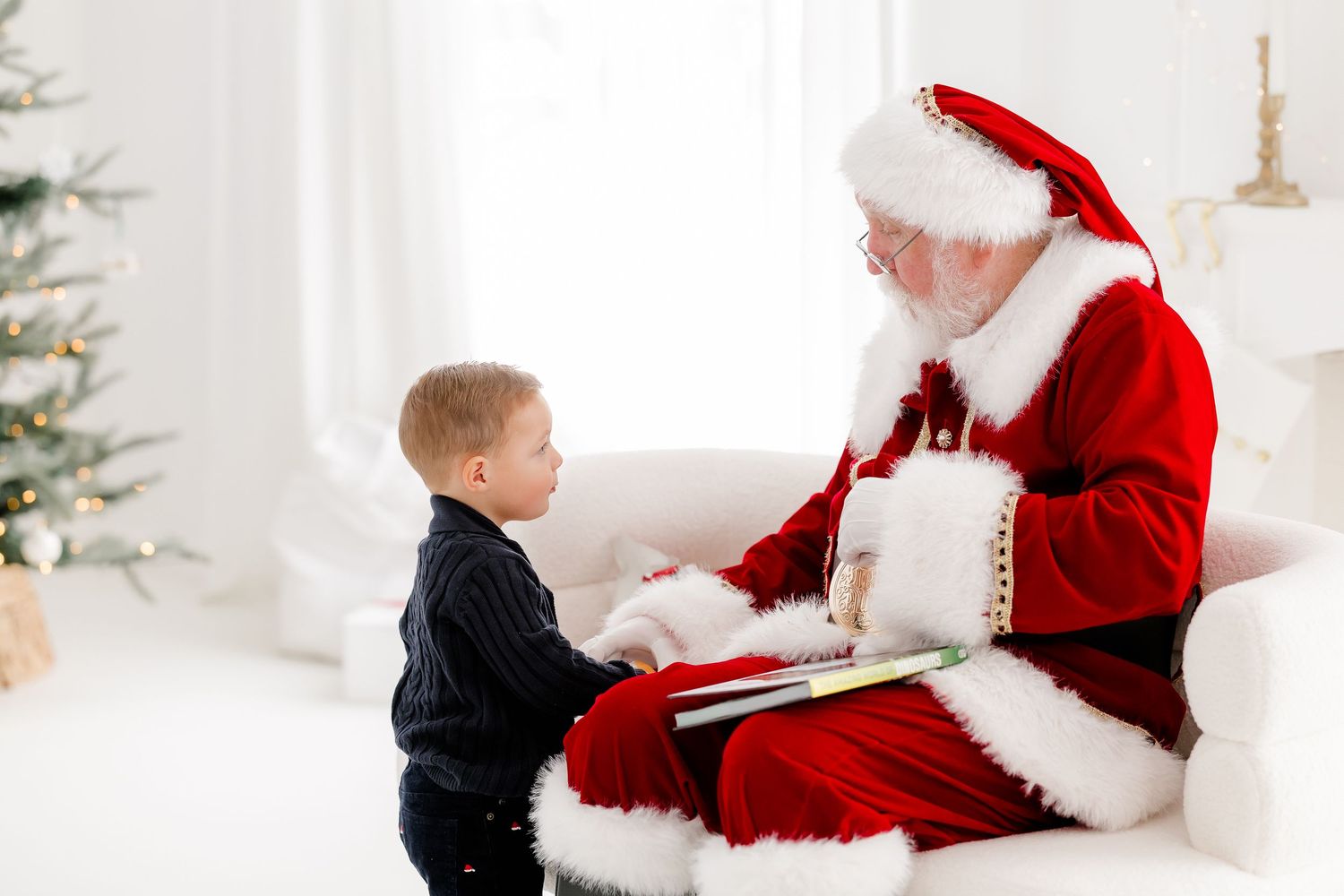 A young boy sits with Santa Claus reading a book together near a Christmas tree in a bright white room.