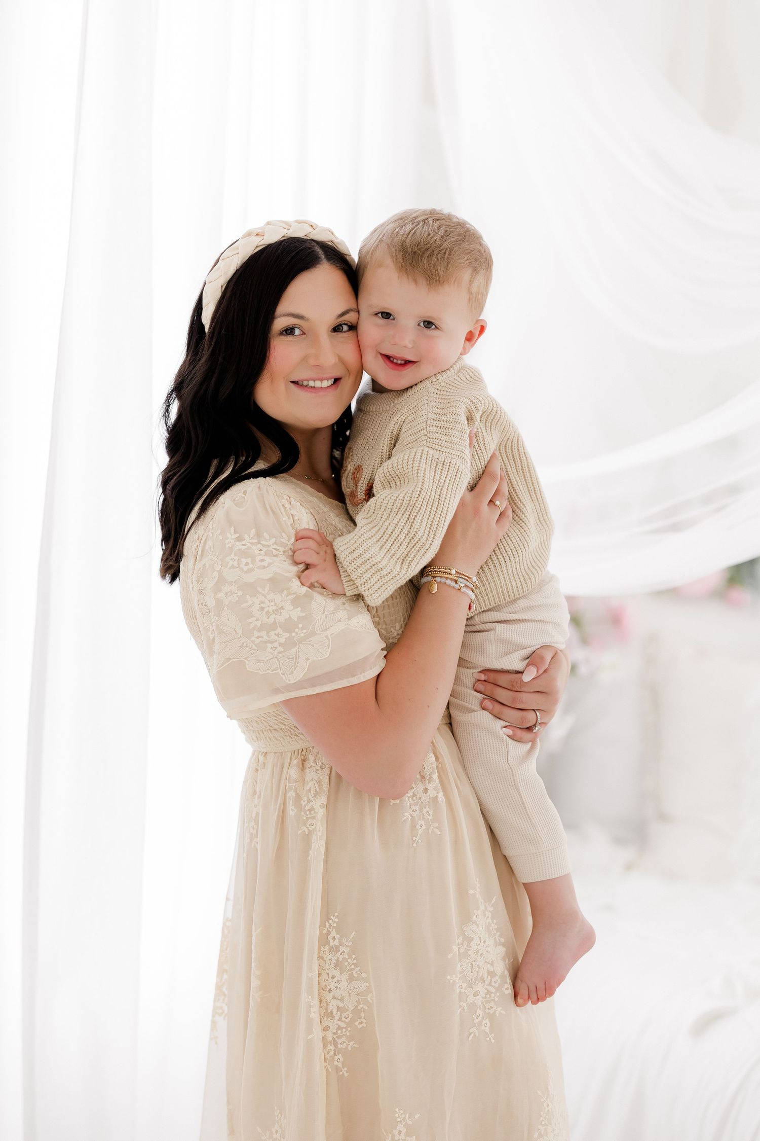 A mother and baby share tender moments in a bright white studio wearing matching cream-colored outfits.
