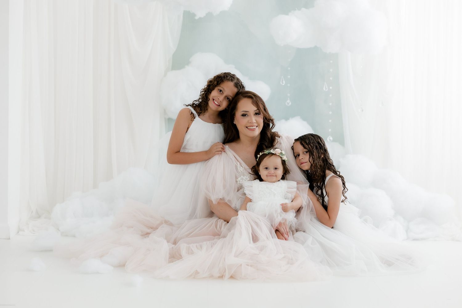 A mother and three daughters in white dresses pose together in a dreamy cloud-themed photo studio setting.