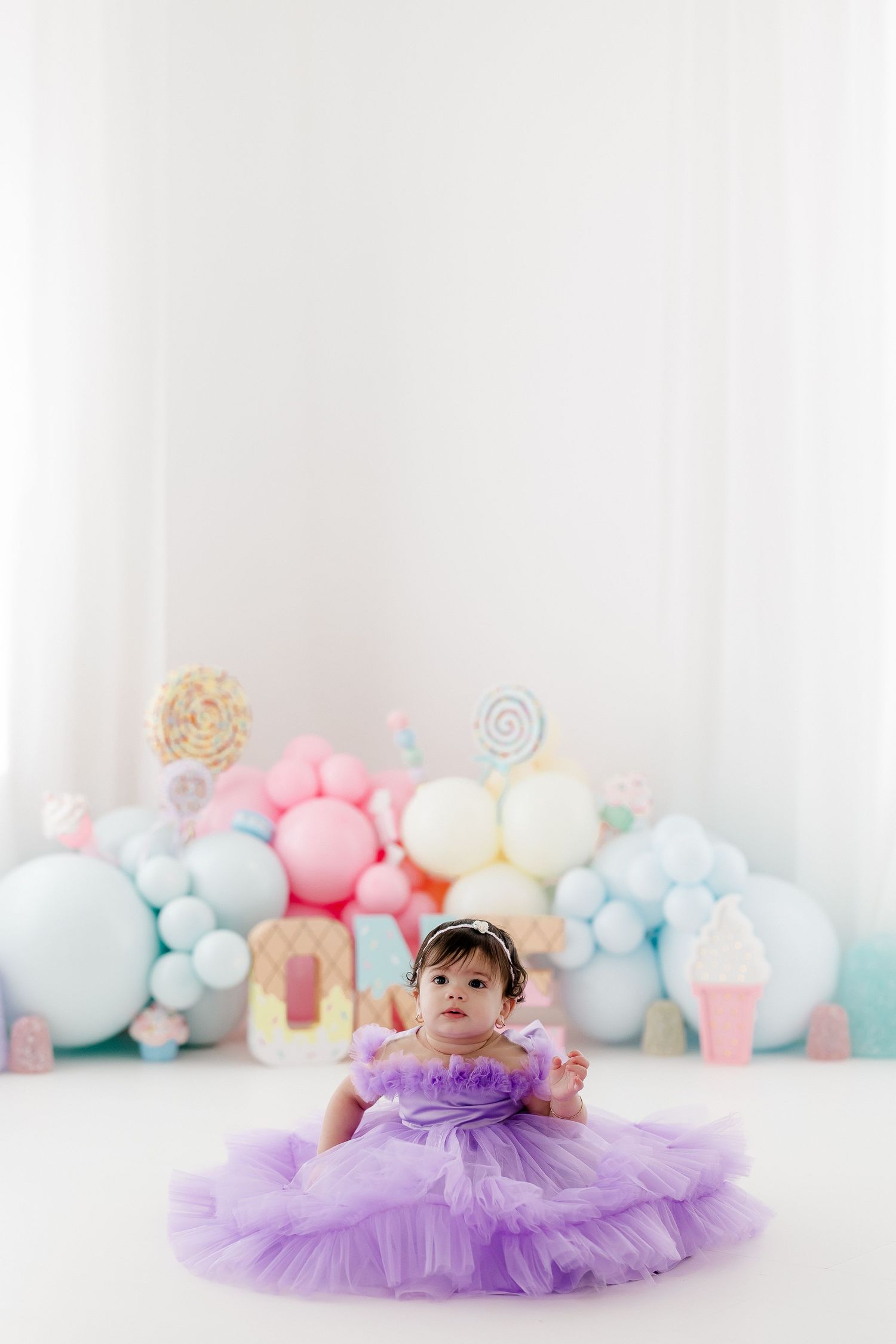 Adorable baby wearing purple tutu dress lying on white floor with pastel balloon arch and decorative backdrop.