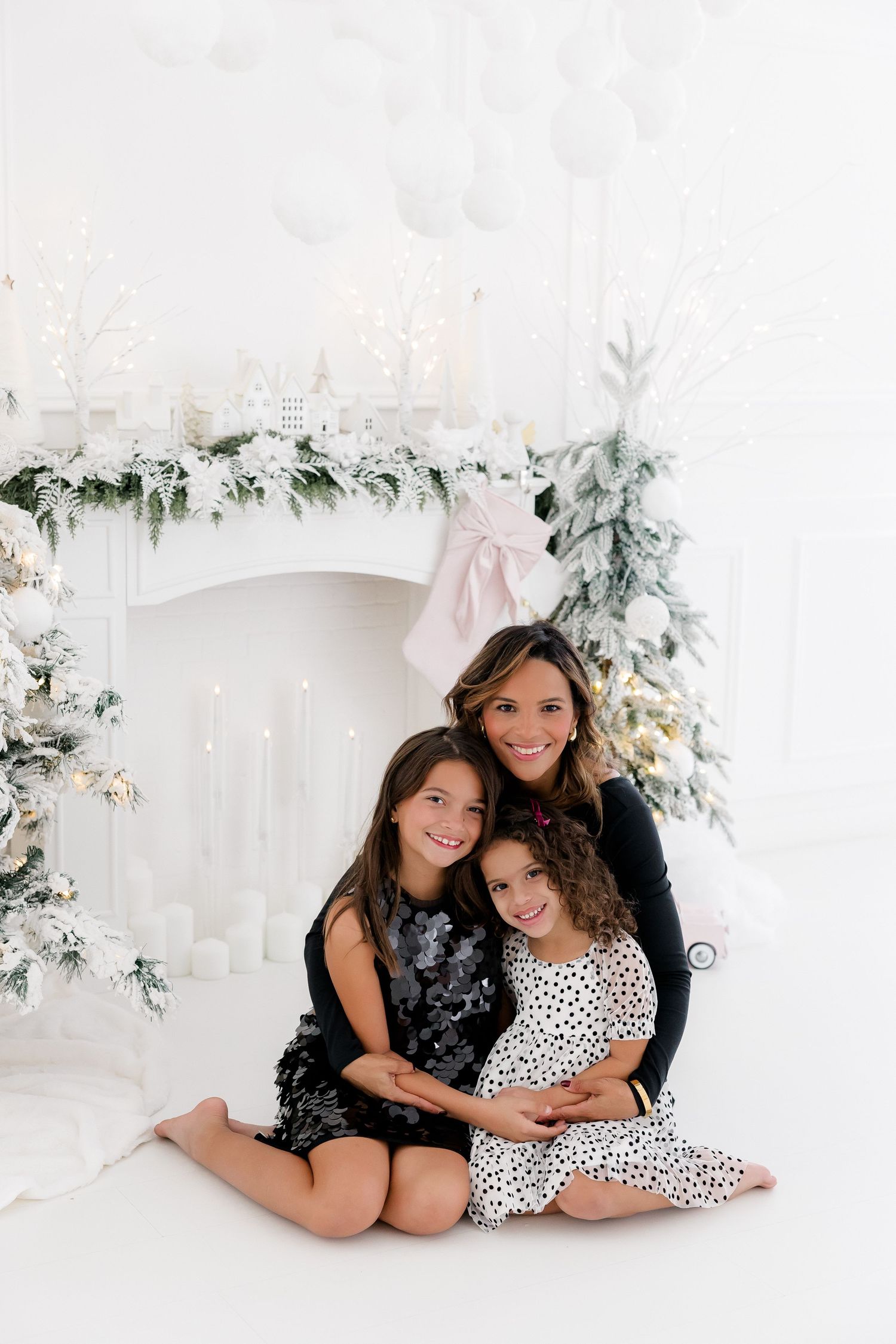 Sisters in holiday attire share sweet moment by Christmas tree in white studio setting.