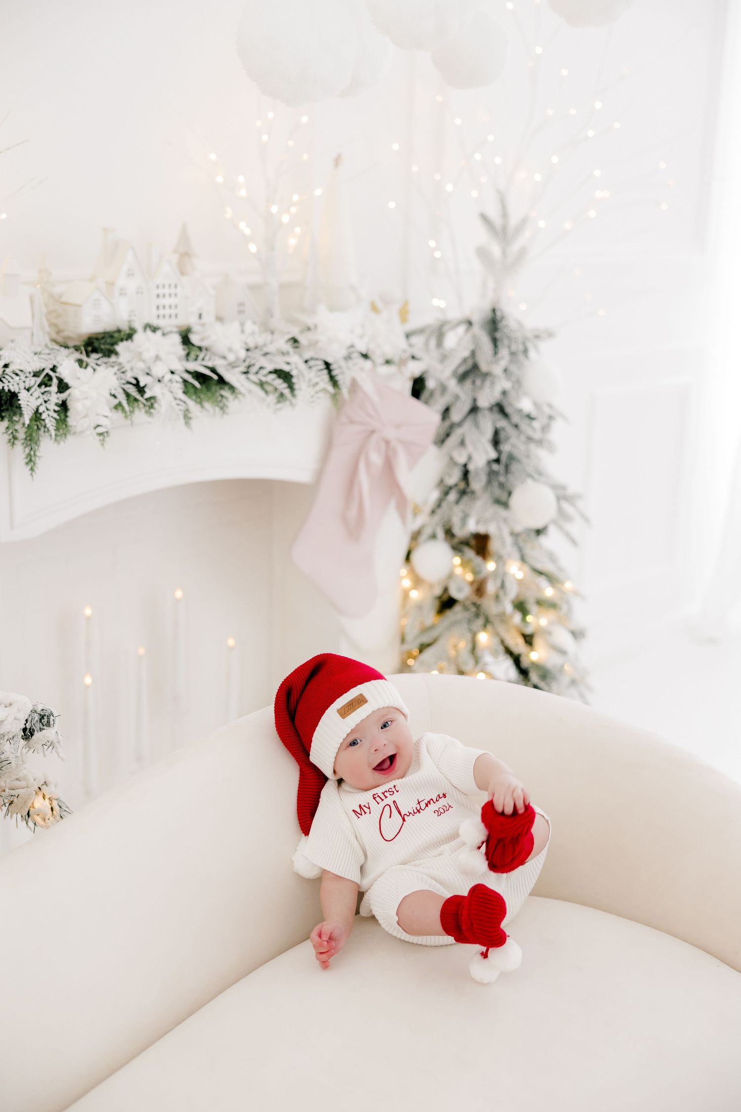 Baby in Santa outfit and red mittens sitting on white couch with holiday decorations.