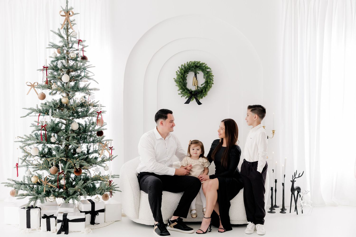 A family shares festive moments together near a decorated Christmas tree in an all-white studio space.