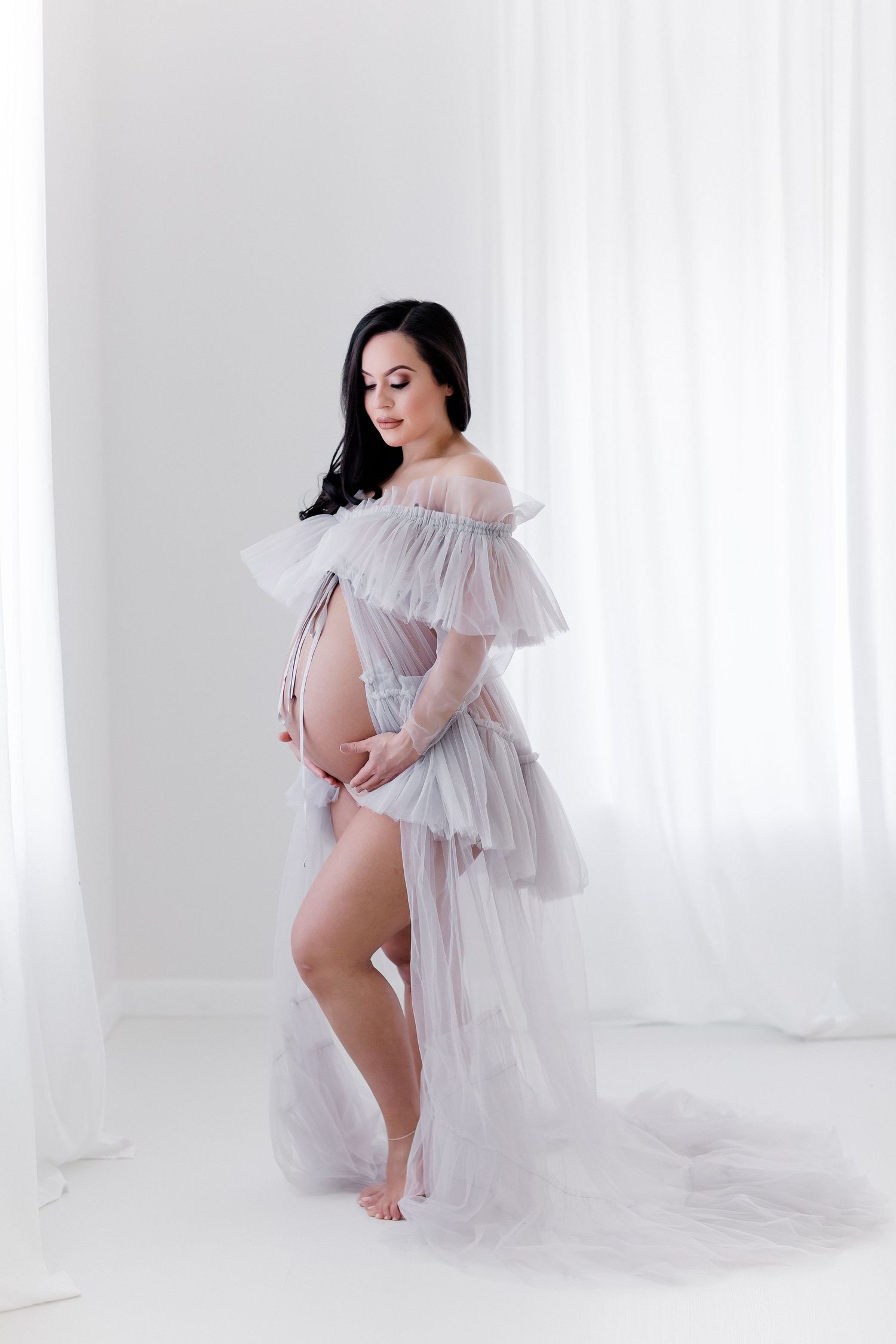 Artistic maternity portrait with flowing white tulle dress against minimalist white backdrop.