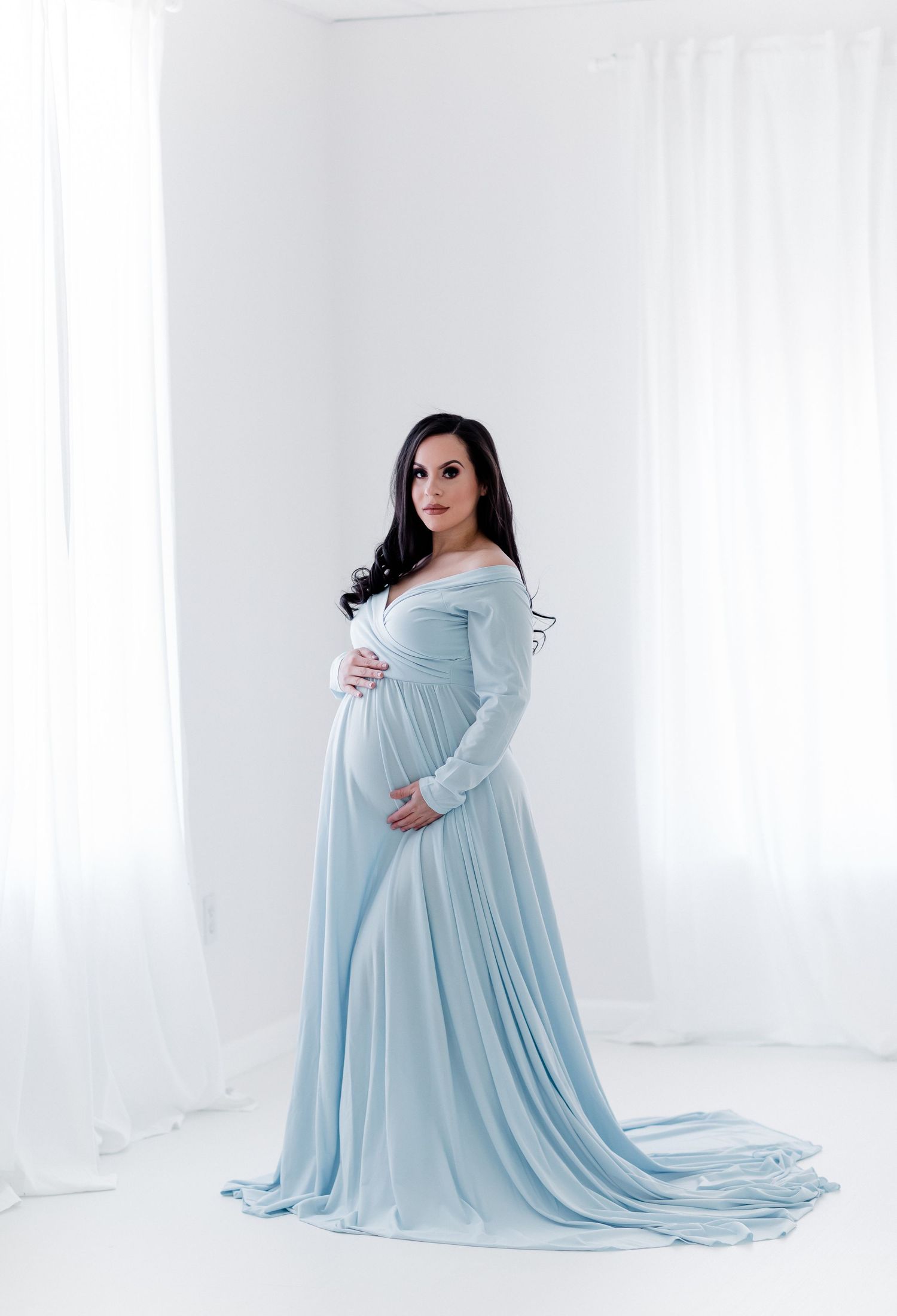 Elegant maternity portrait in flowing pale blue gown with long train in minimalist white setting.