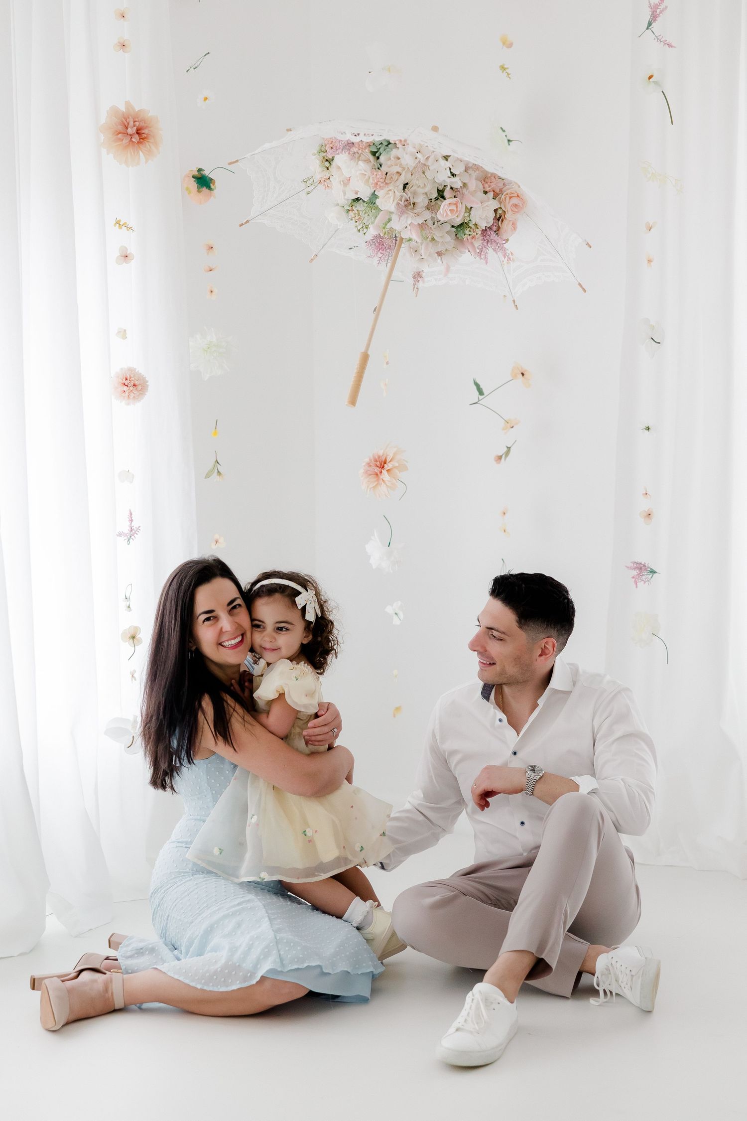 A family in light pastel outfits laughs and plays together during a bright white studio photoshoot.