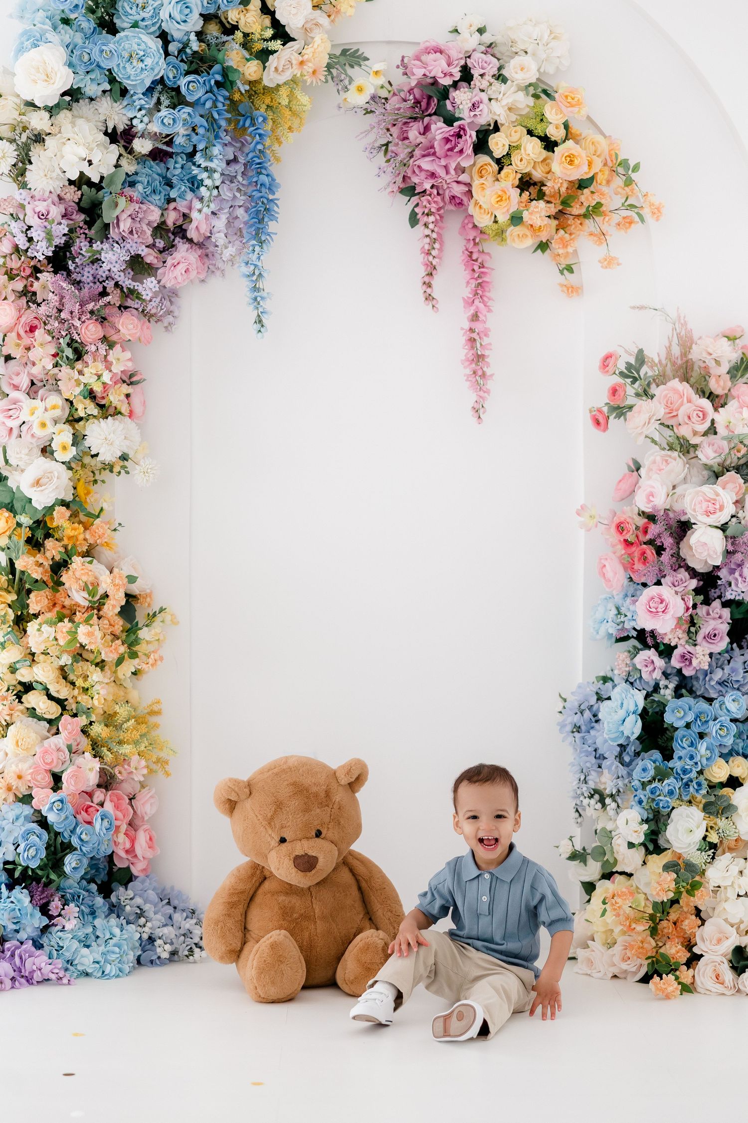Beautiful floral arch with pink, blue and peach flowers frames a small child sitting next to a large teddy bear.