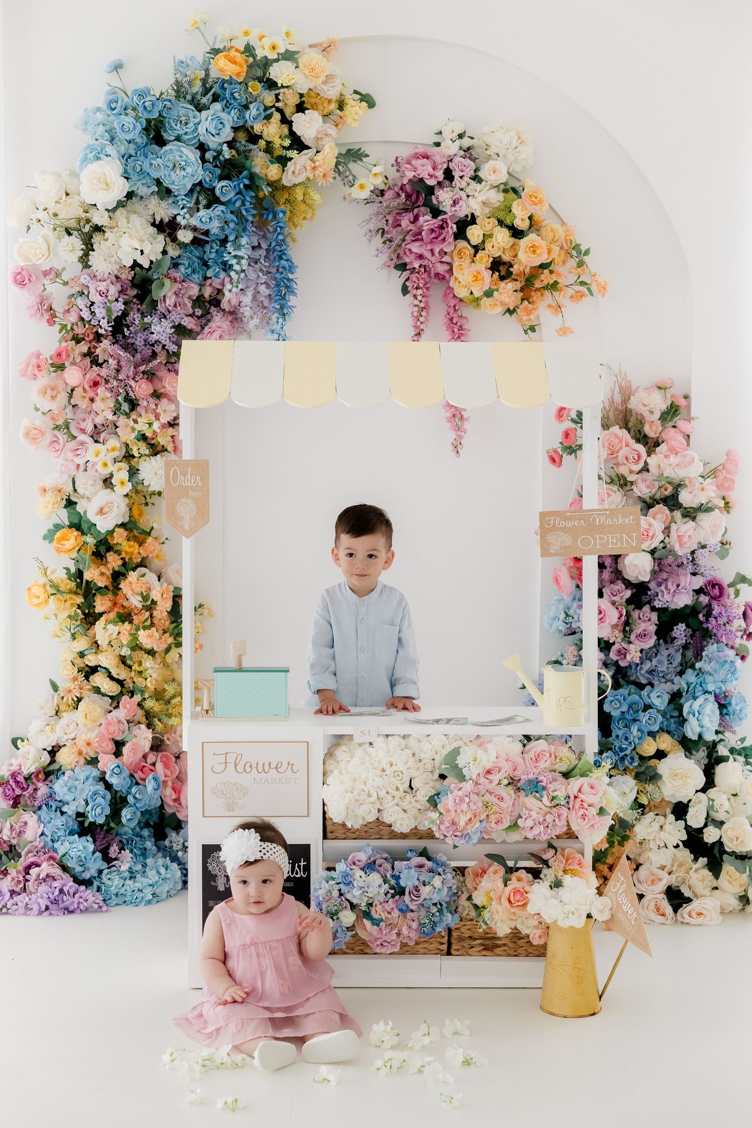 Beautiful floral studio setup with pastel pink, blue, and yellow flowers adorning white arched backdrop for photography session.