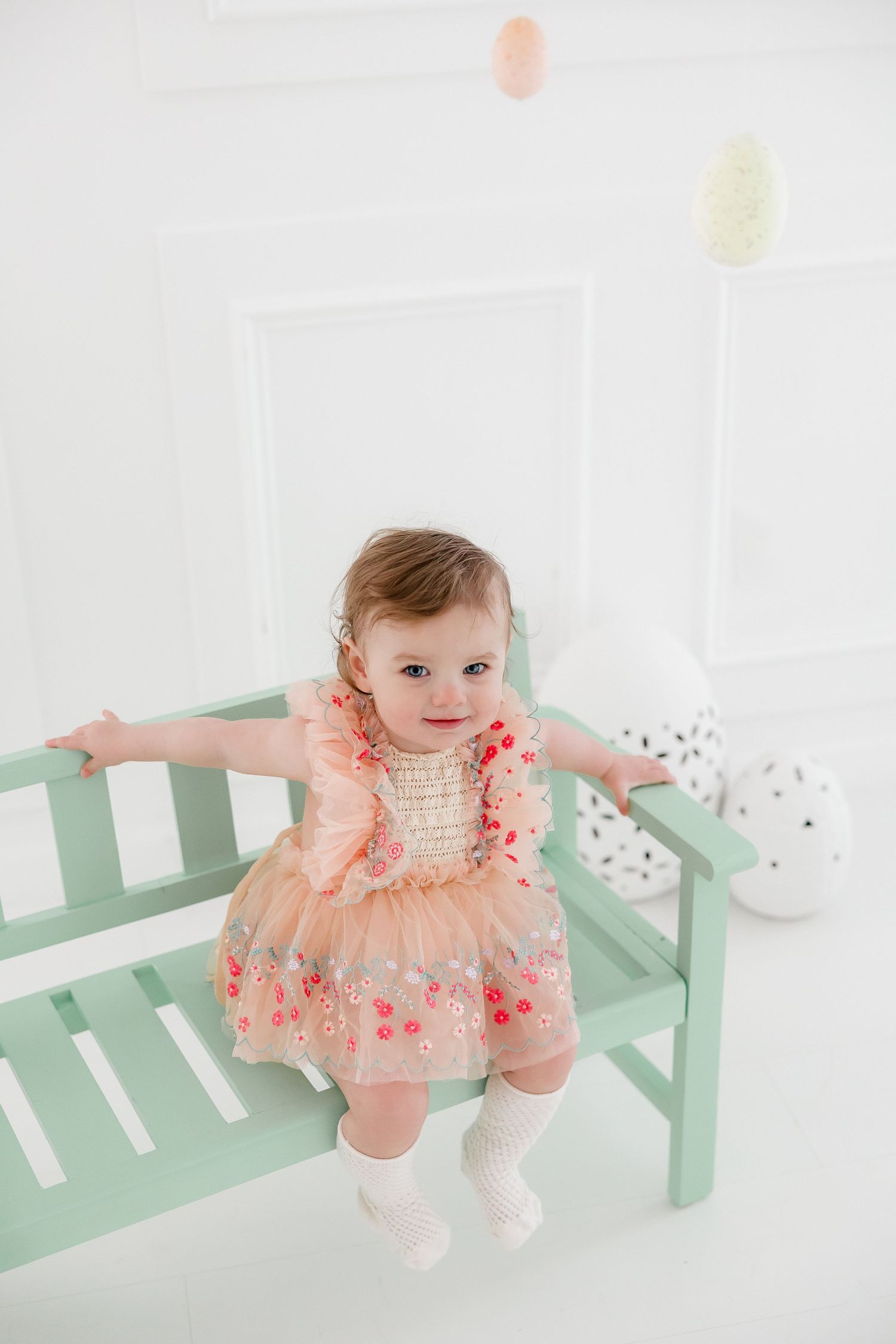 A baby in a peach-colored tulle dress sits on a mint green bench in a bright white studio setting.
