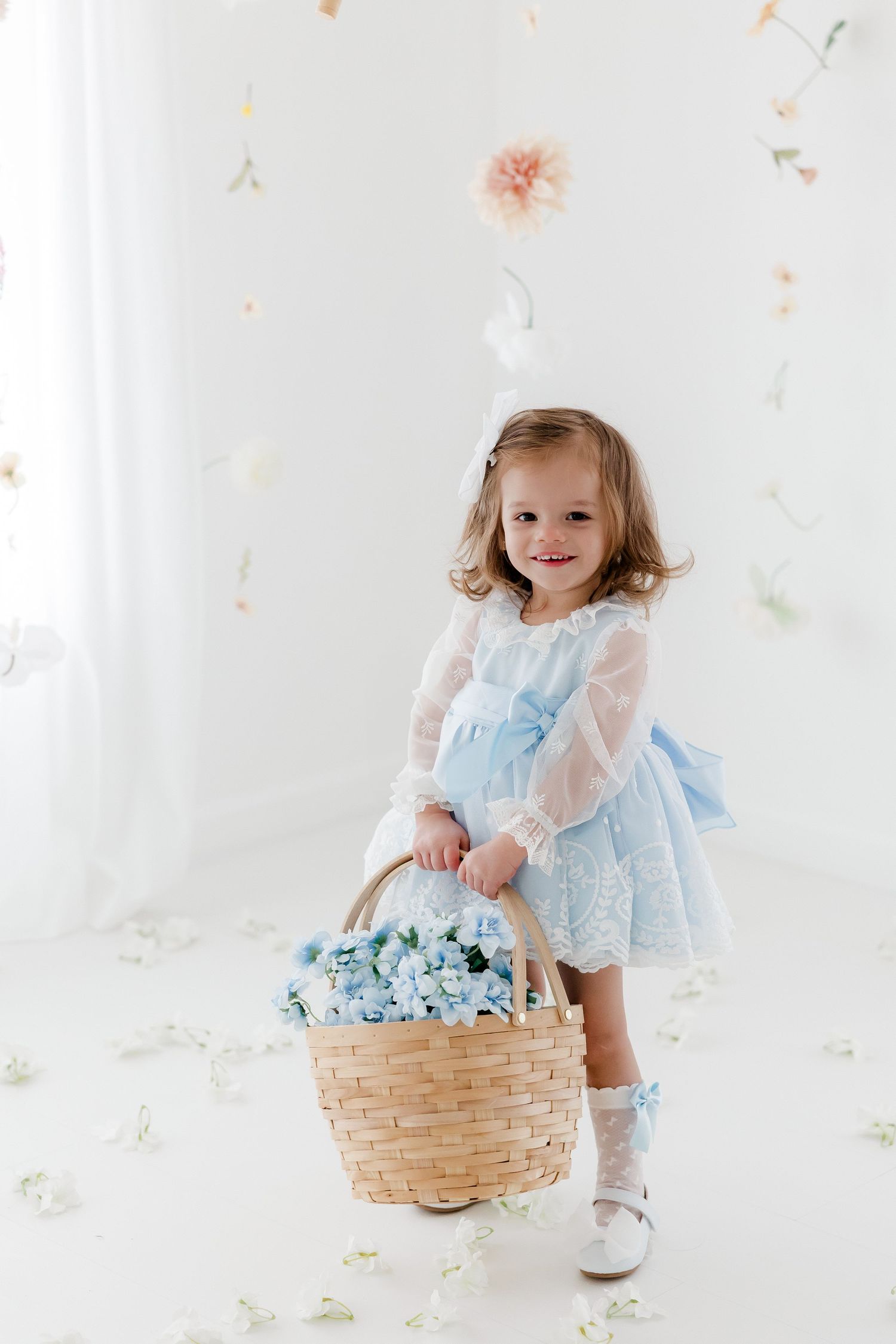 A child in a light blue dress holds a woven basket filled with blue flowers against a white floral backdrop.