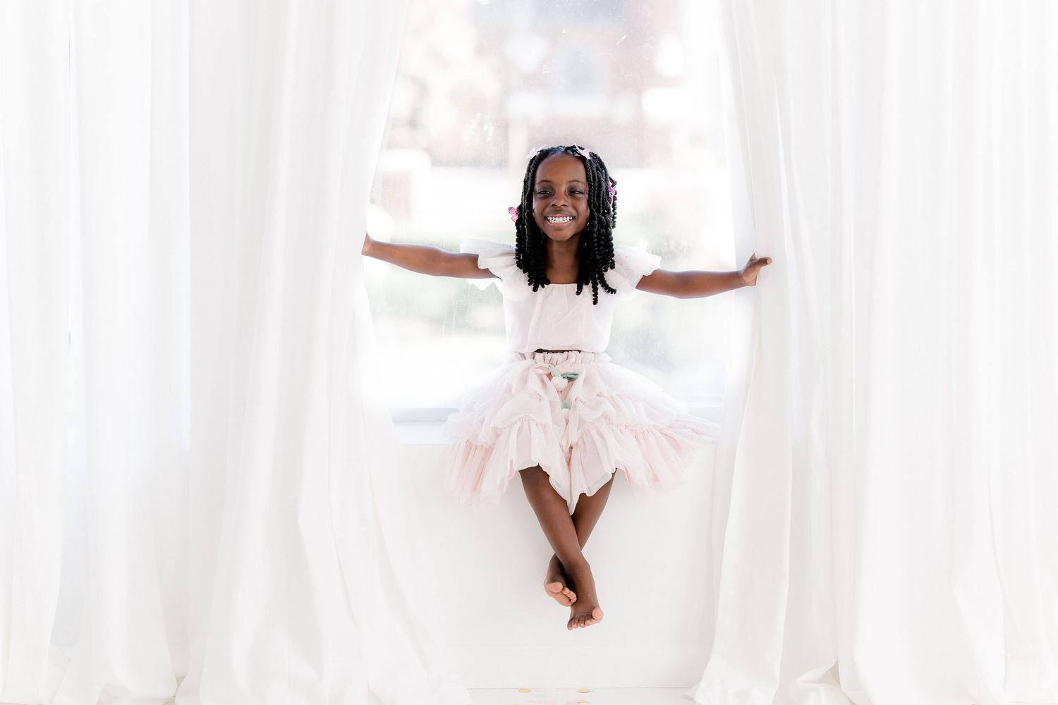 Child in white dress jumping playfully against a bright white curtain background.