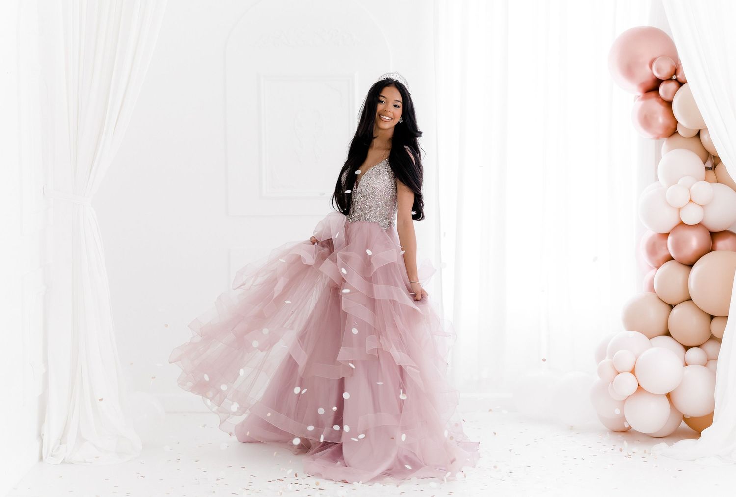 A dreamy pink tulle gown flows gracefully in a maternity photoshoot setting with soft pastel balloon decorations.
