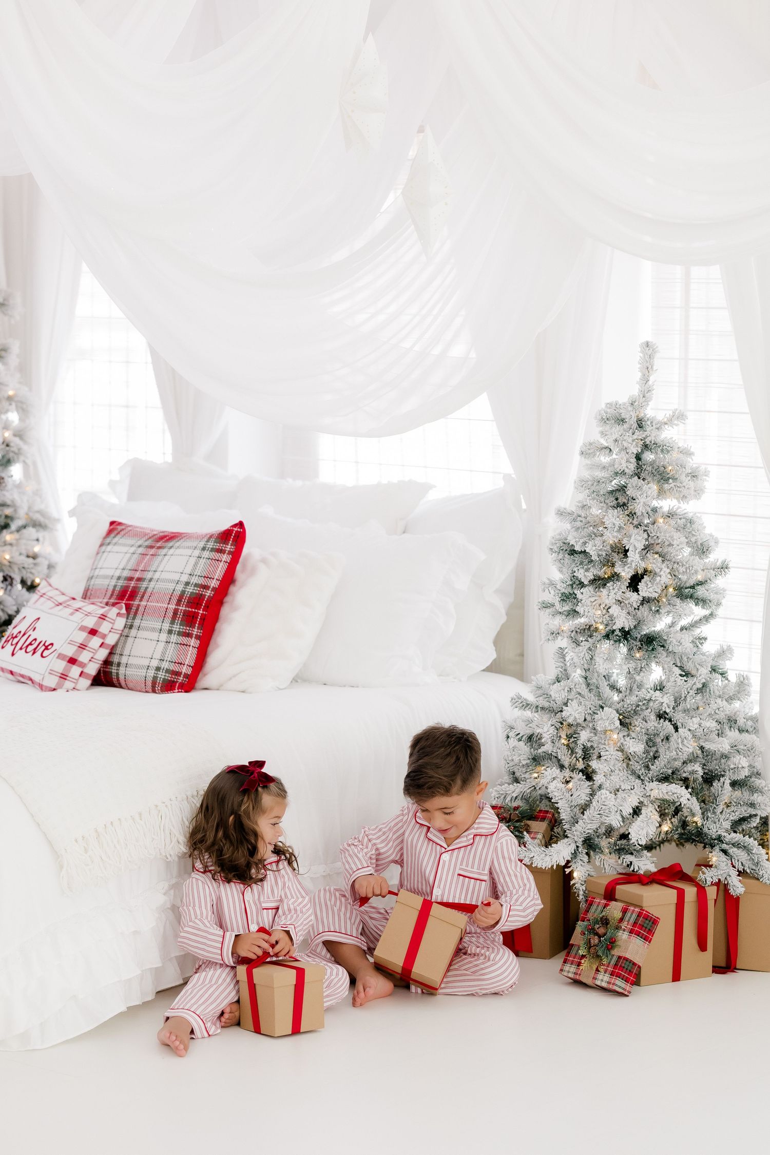 Two children in red striped pajamas open Christmas gifts beside a flocked tree in a white decorated bedroom.